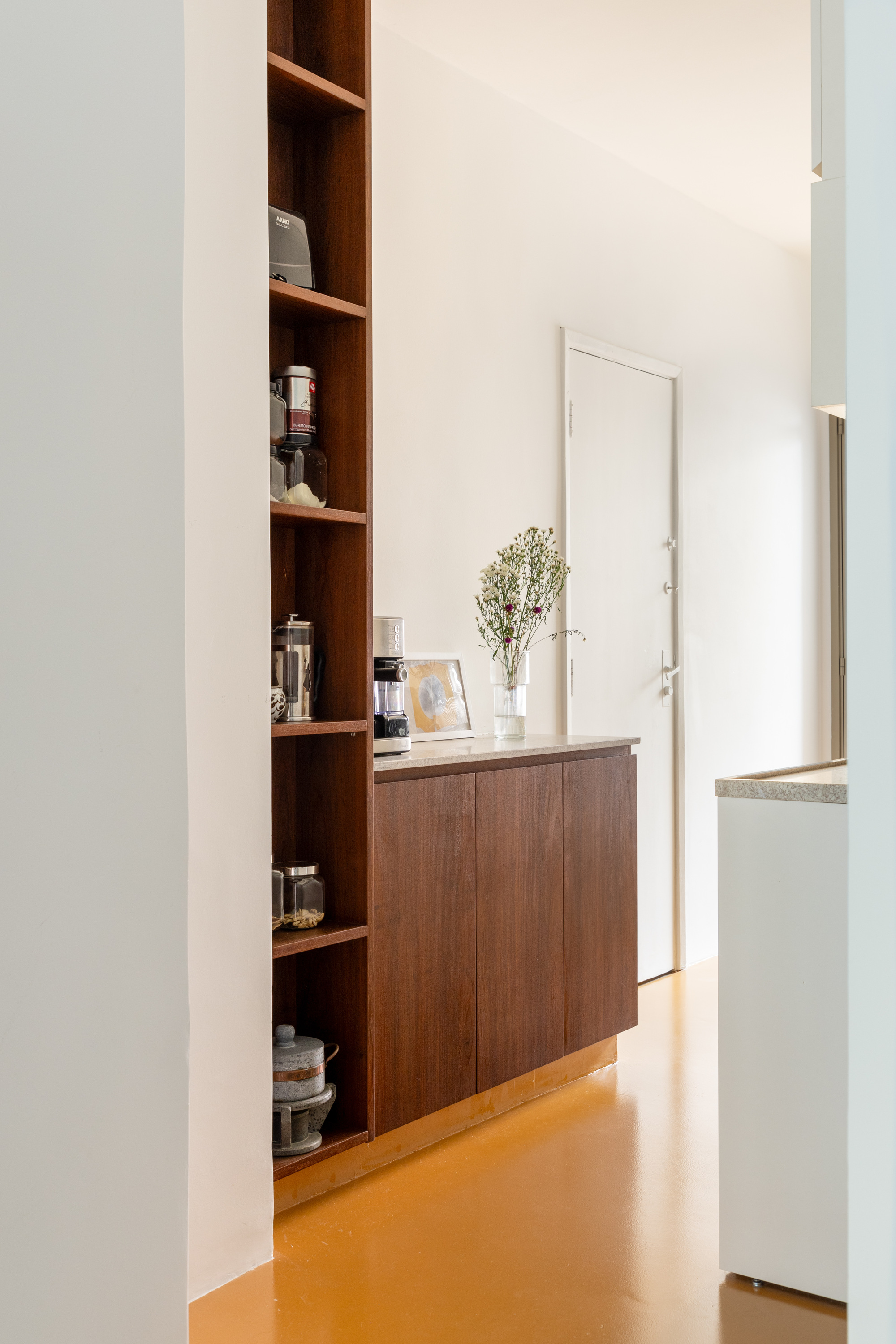 Walnut cabinet and tall open shelving holding coffee gear, with a vase of dried flowers beside a white door