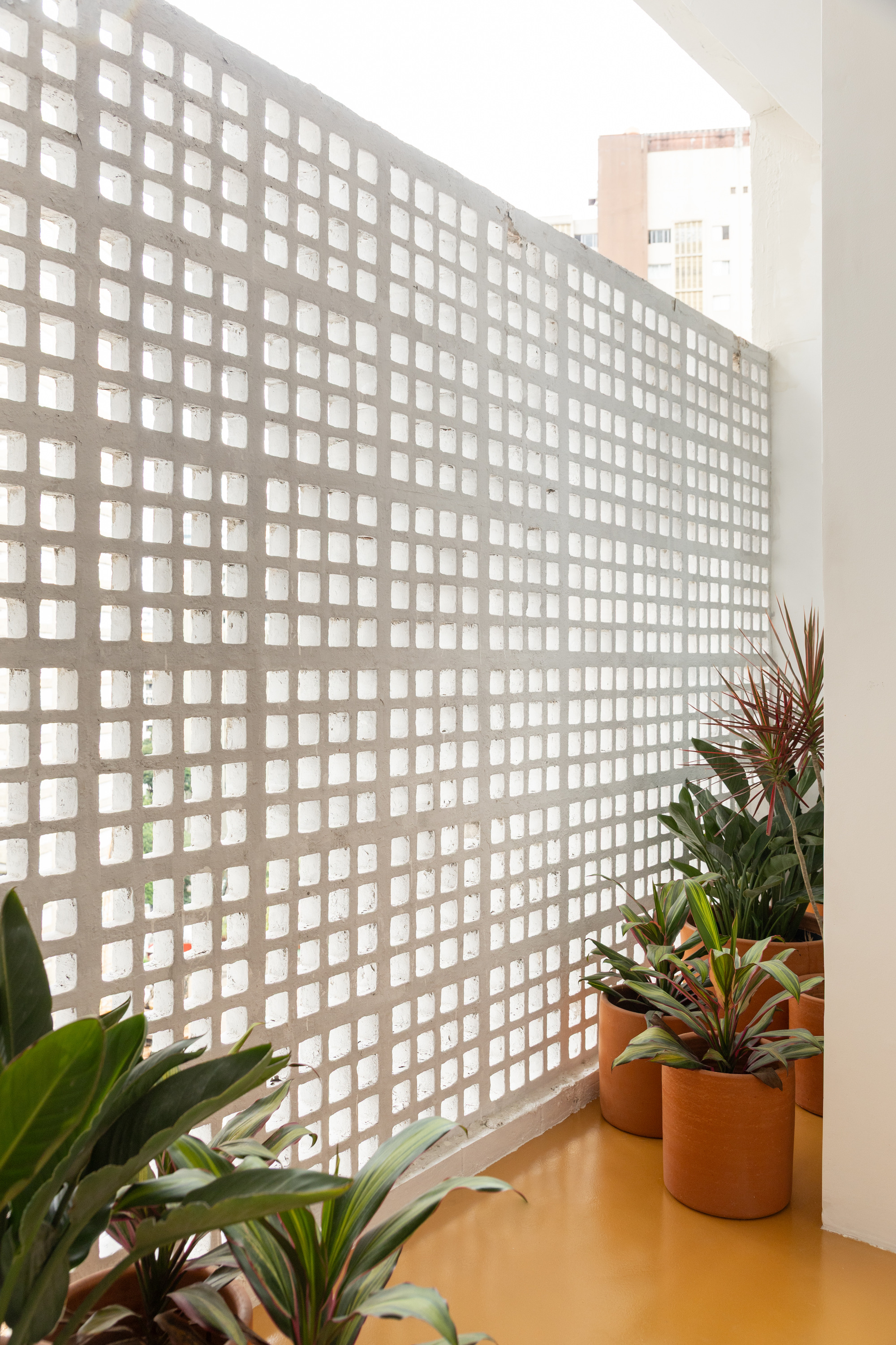 Perforated white concrete block screen along a balcony with potted tropical plants on a yellow floor, a neighboring tower beyond