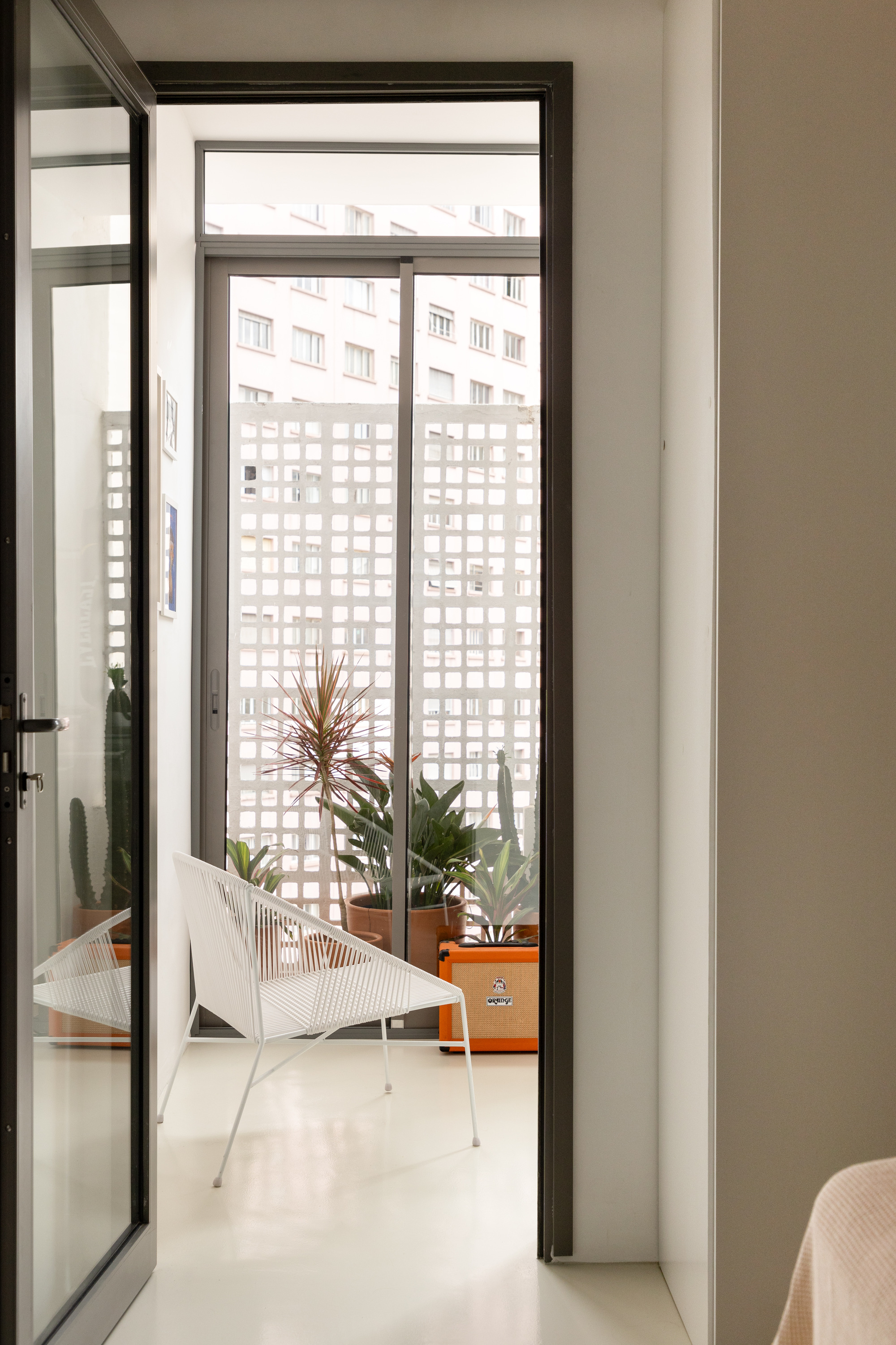Doorway view to a balcony with a white wire chair, potted plants and a lattice screen facing an apartment block