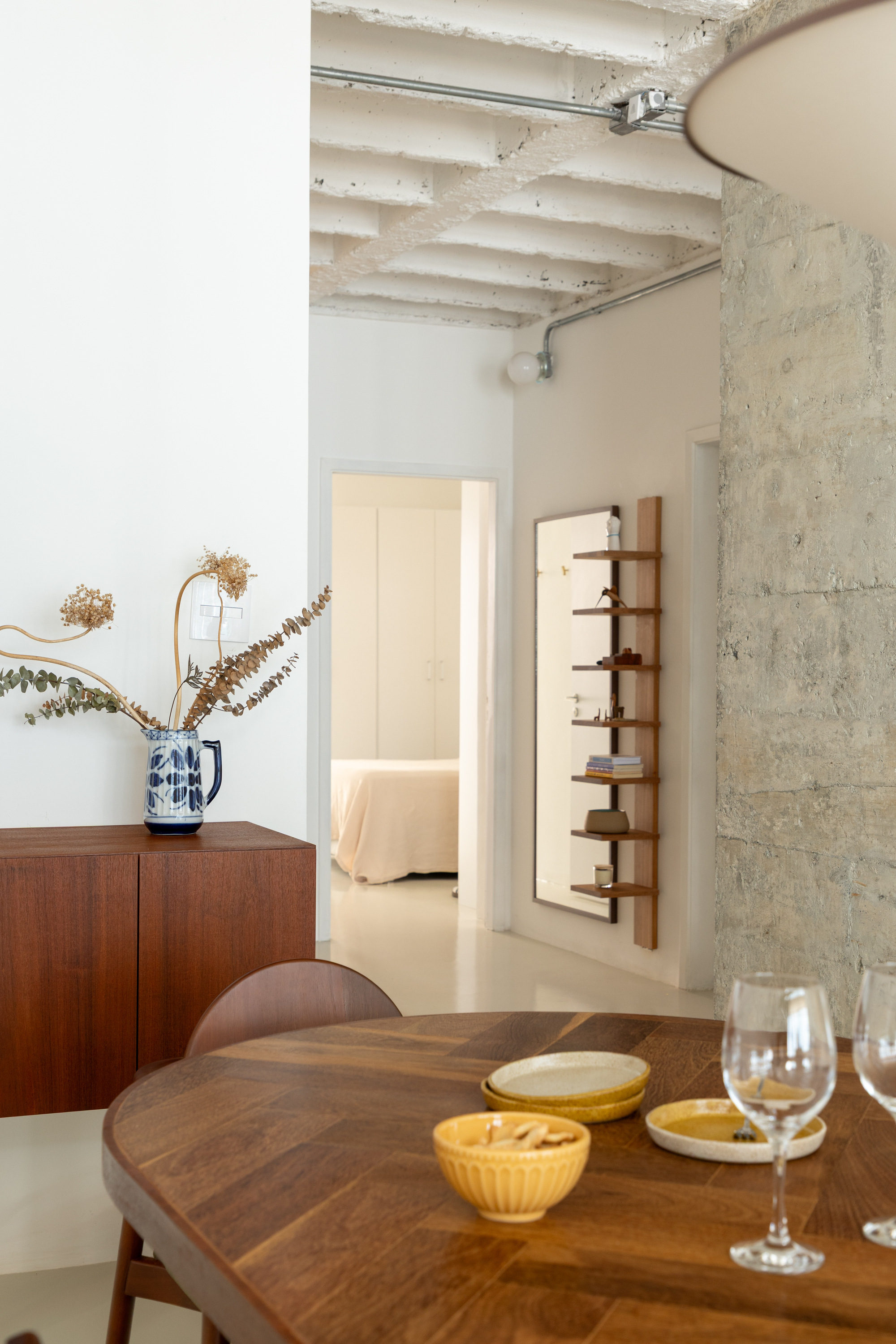 Round wooden dining table with yellow ceramic bowls and wine glasses beside a dried-stem vase on a teak sideboard, soft daylight