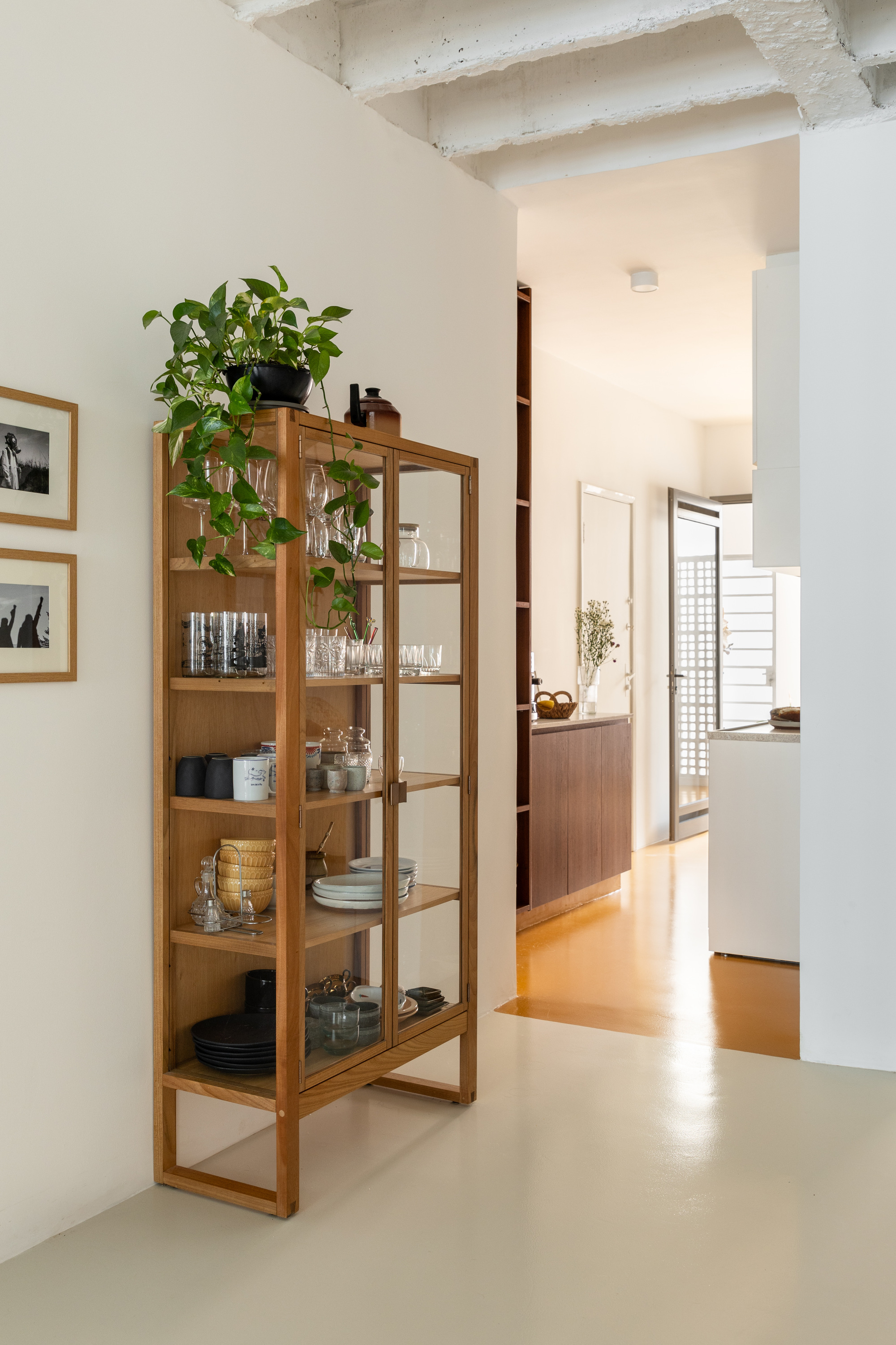 Glass-fronted timber display cabinet holding glassware and plates, topped with a trailing pothos plant beside a doorway to the kitchen