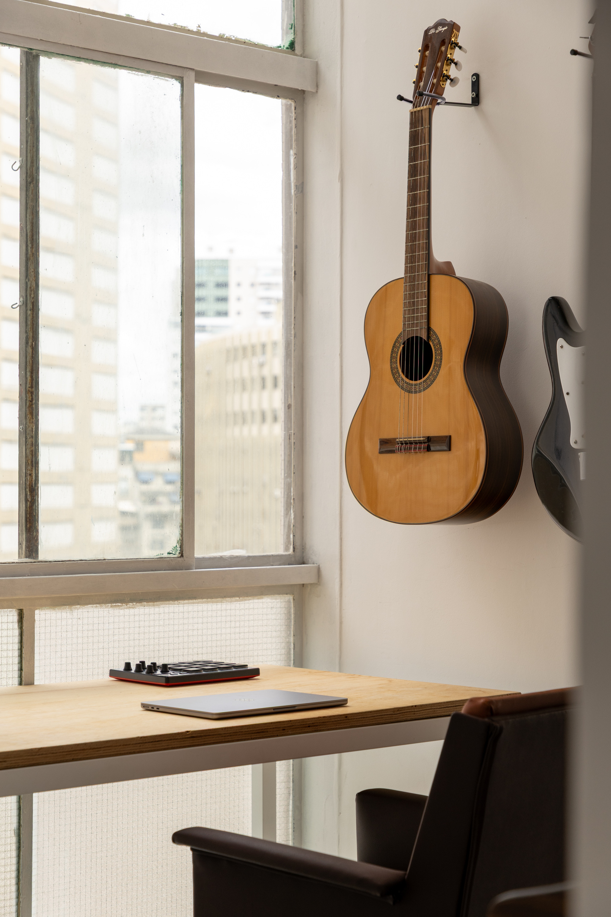 Classical guitar hanging on a white wall above a plywood desk and leather chair, with a tall window showing neighboring towers