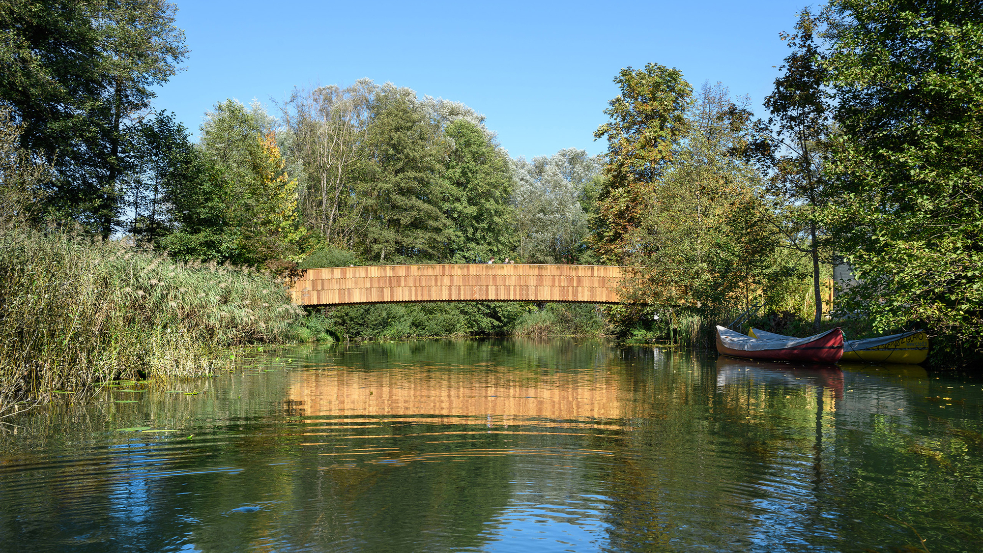 Timber footbridge spanning a calm river, its shingled side reflected in the water beside moored canoes in clear sun