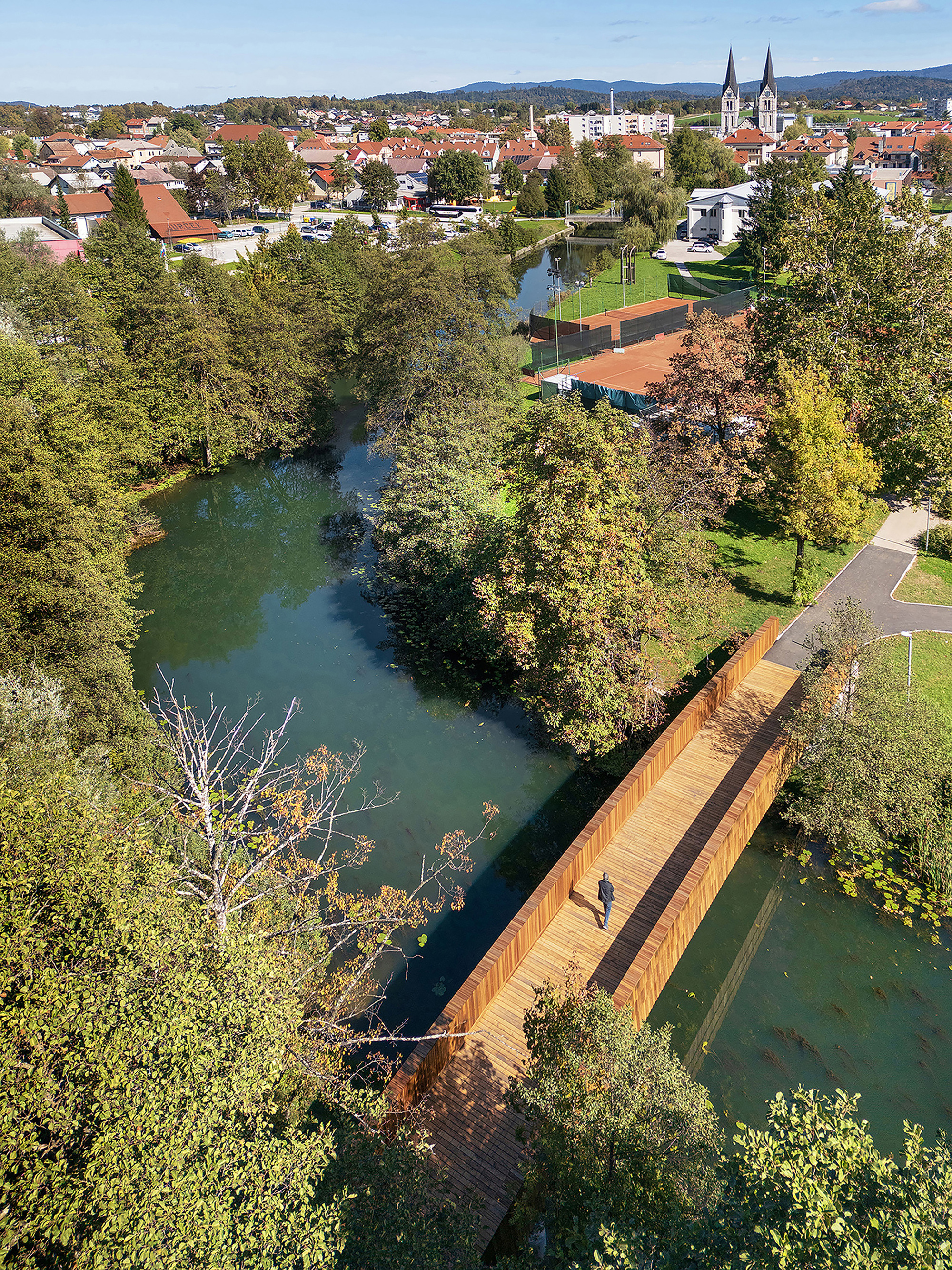 Aerial view of a curved timber footbridge crossing a green river, with a lone pedestrian and a town beyond
