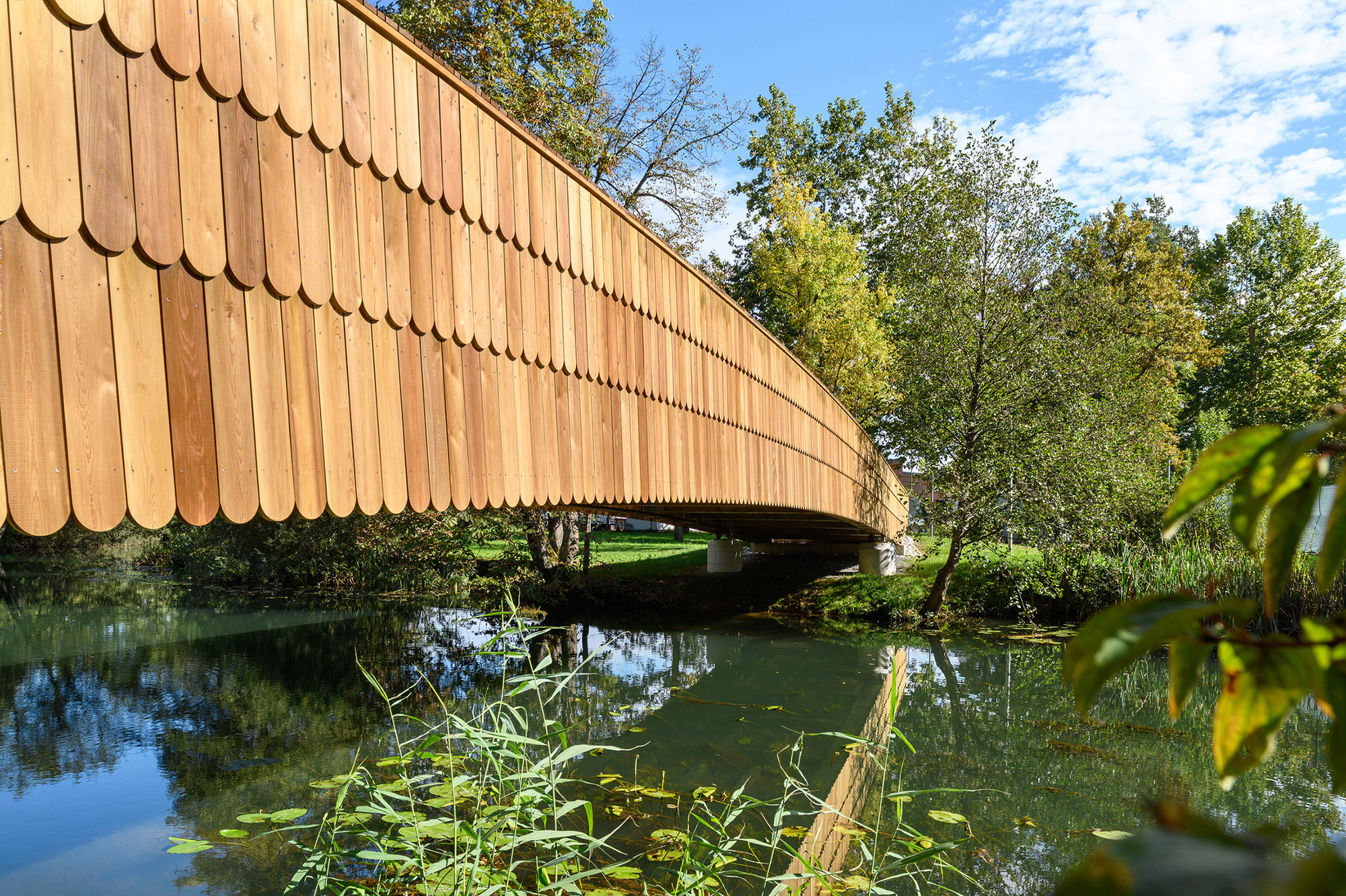 Side of the bridge clad in rounded wooden shingles, curving over still water under a partly cloudy autumn sky