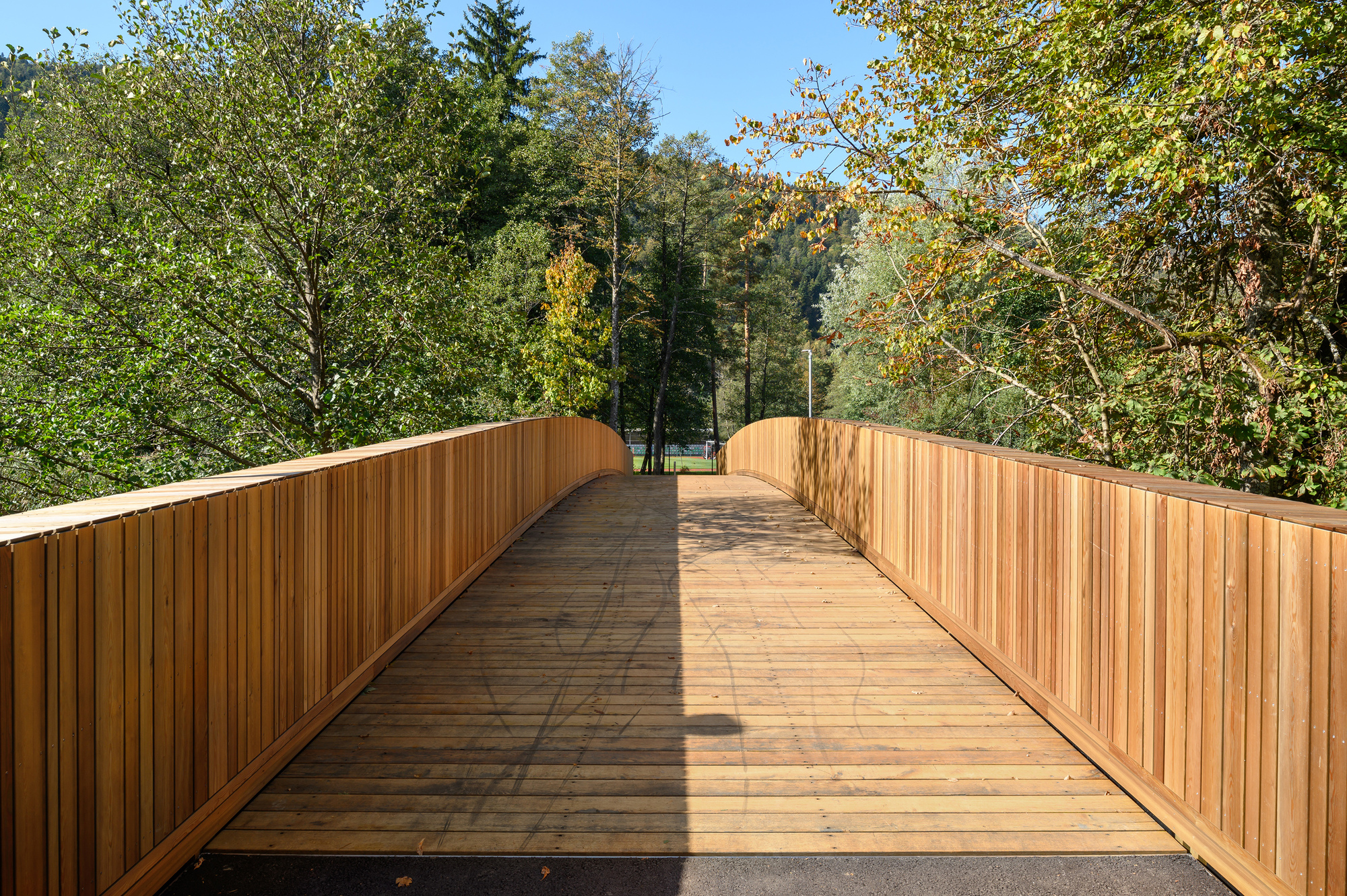 Deck of the footbridge with slatted timber boards and vertical plank parapets, dappled by shadows from surrounding autumn trees