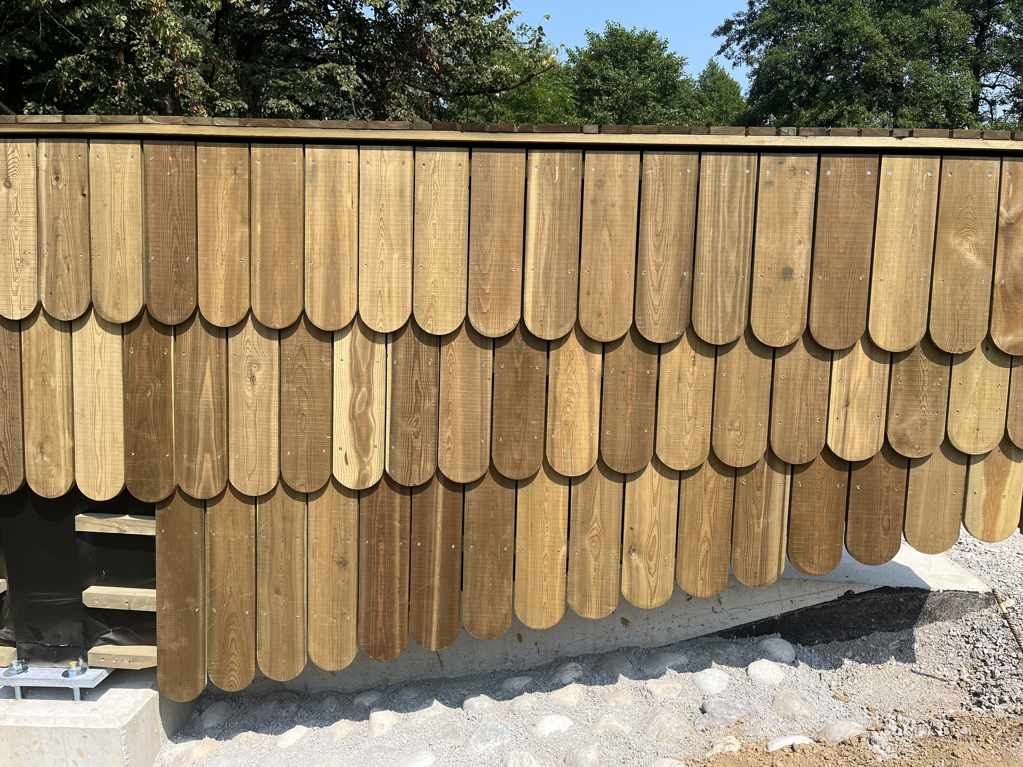 Close-up of overlapping rounded timber shingles on the bridge abutment, resting on a concrete footing and gravel in bright sun