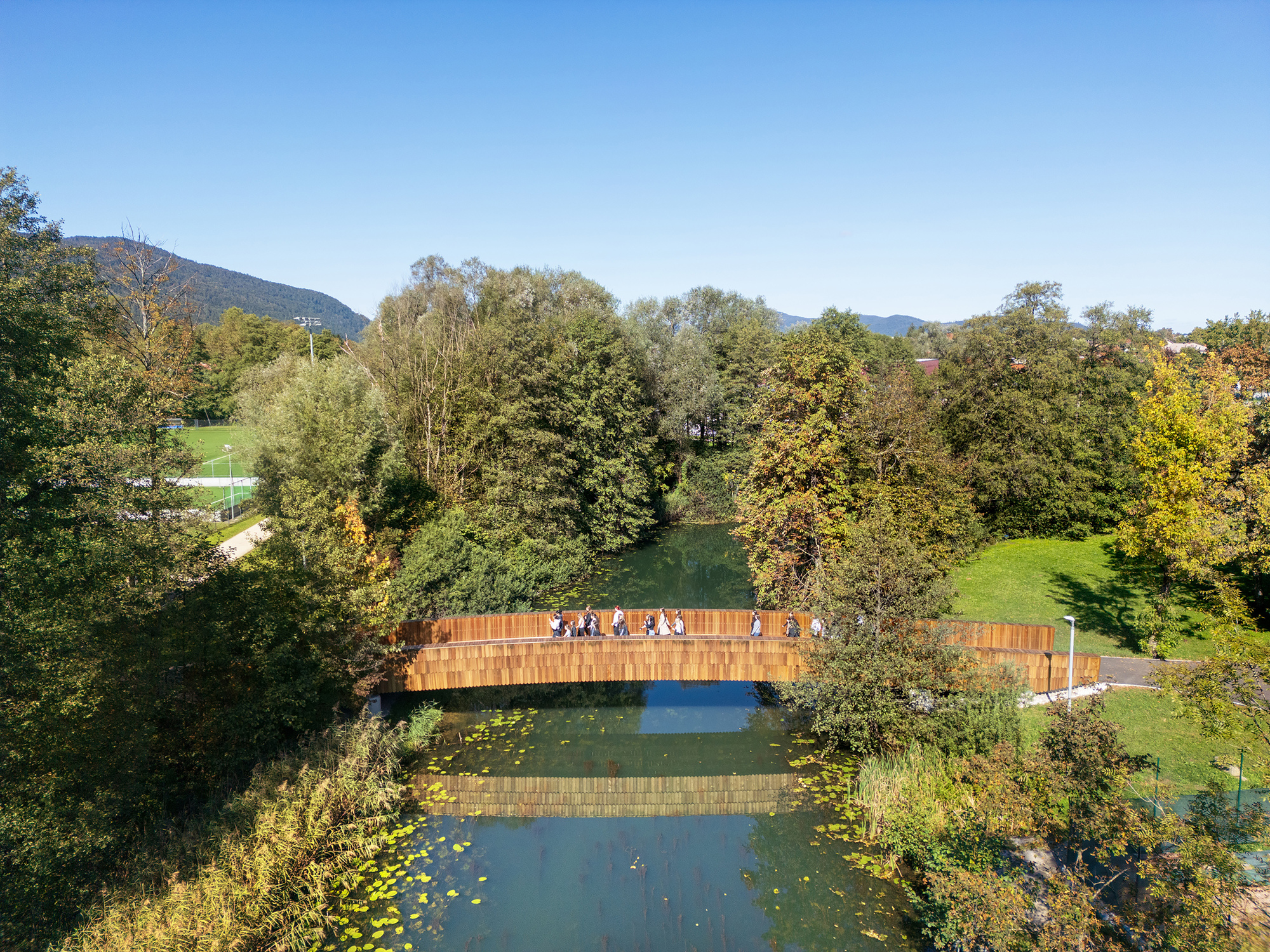 Drone view of the arched wooden footbridge over a green river, with a few pedestrians crossing and water lilies below