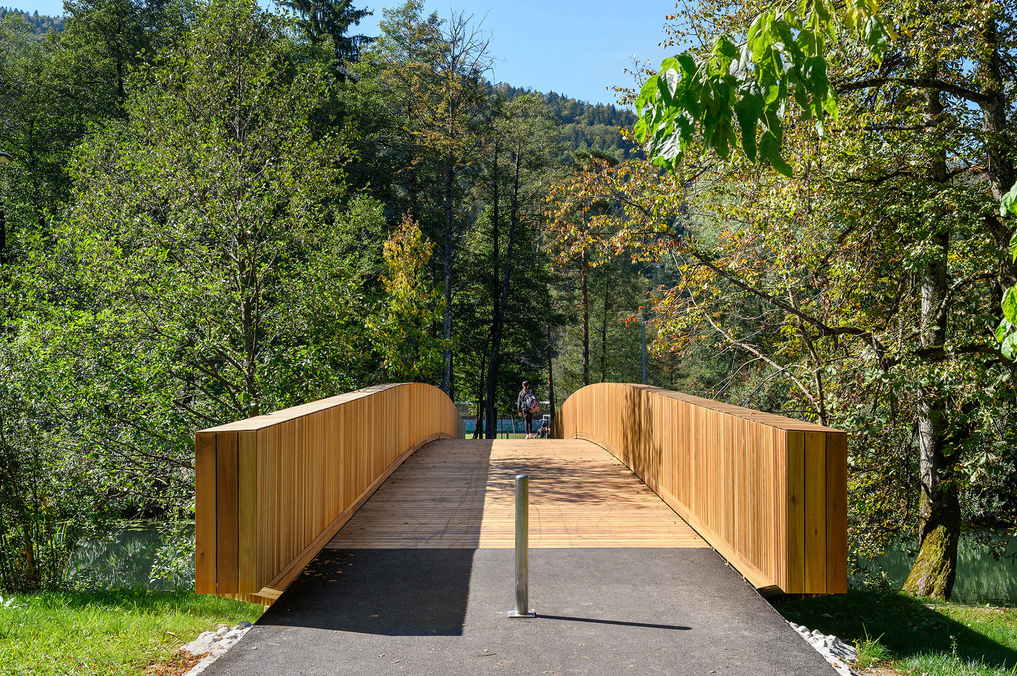 Head-on view of the bridge deck and tall cedar parapets, with a steel bollard on the asphalt path and a walker beyond