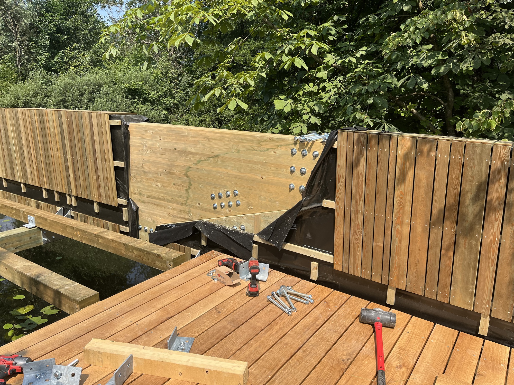 Construction detail of a timber bridge deck with bolted steel plate connection, black membrane and vertical slat cladding under summer foliage