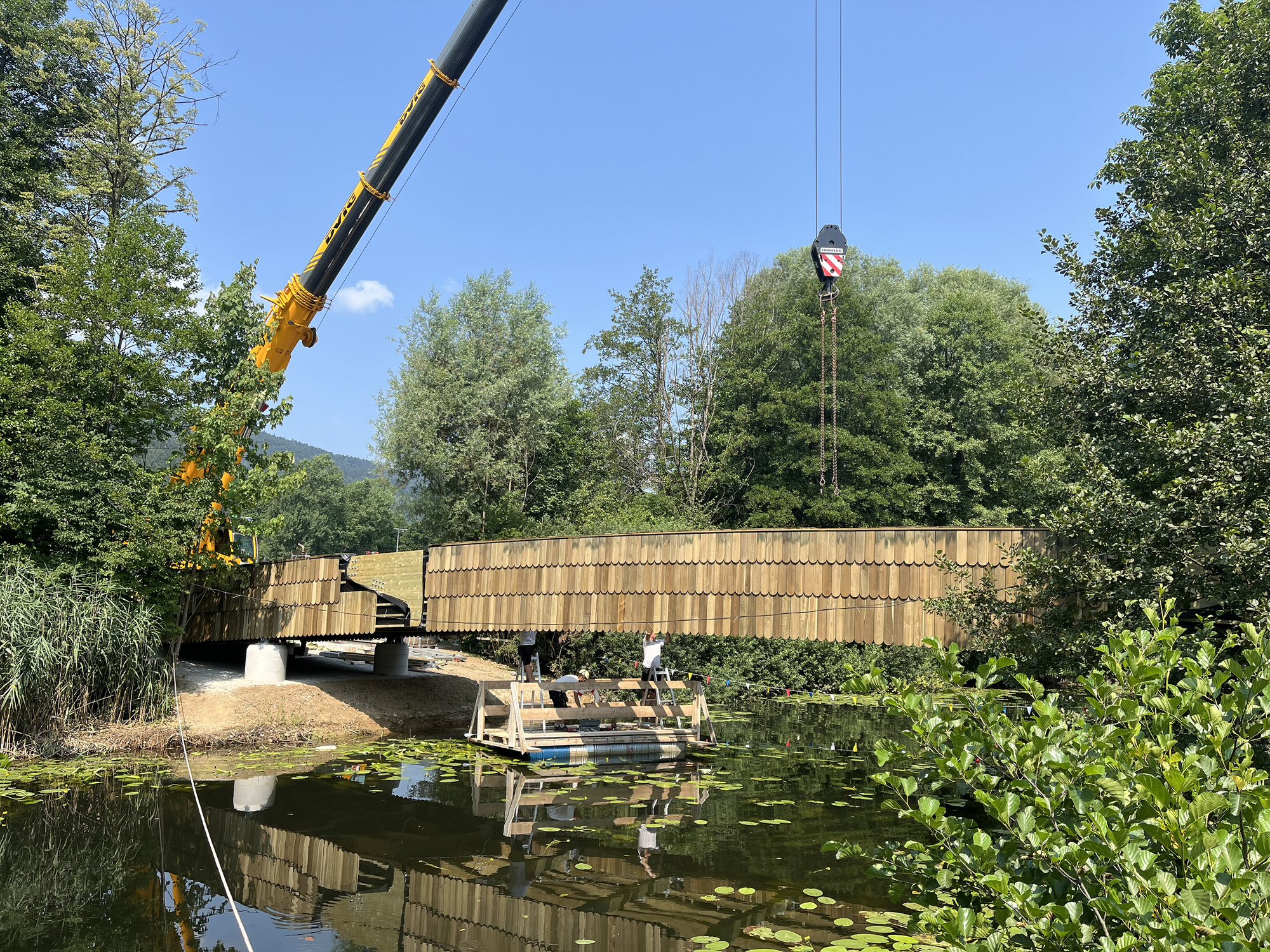 Mobile crane lifting the curved timber-clad footbridge into place over a pond with lily pads on a clear sunny day