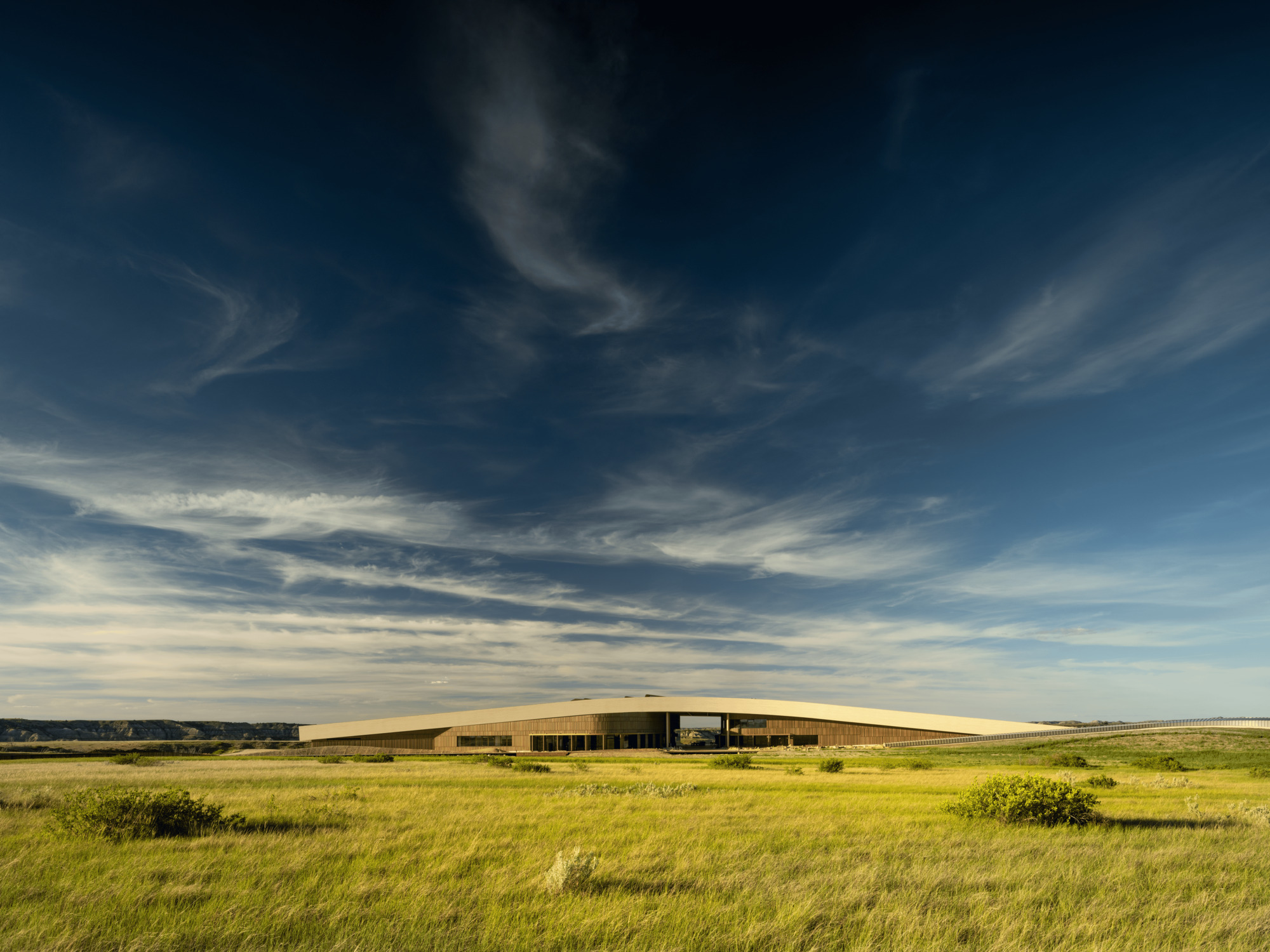 Low, long curved roof of the library building across open prairie grass under streaked clouds at late afternoon