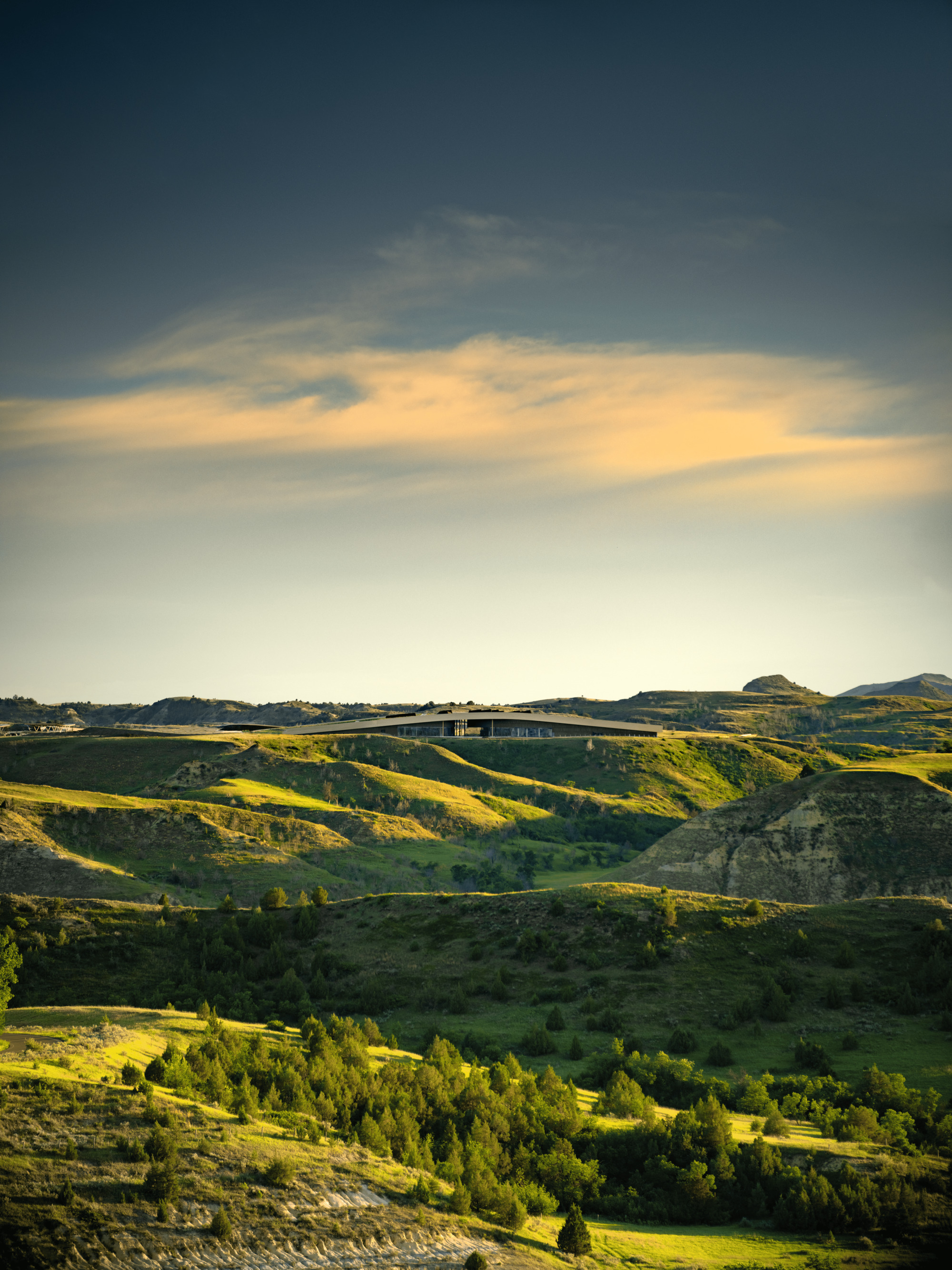 Distant view of the low roofline on a ridge above green badlands hills, lit by a glowing orange cloud at dusk