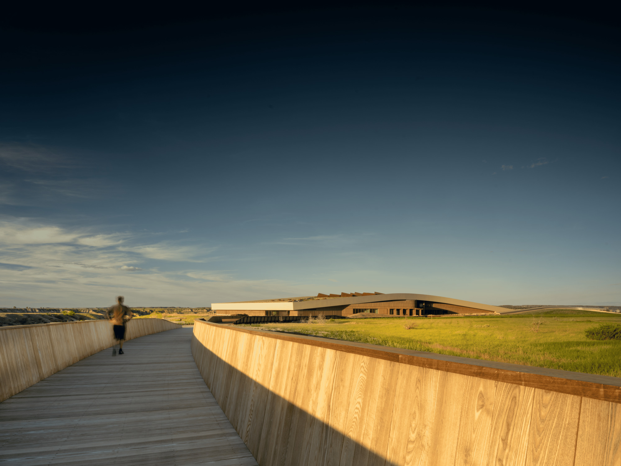 Curving timber boardwalk with tall wooden side wall leading toward the building as a runner passes in evening light