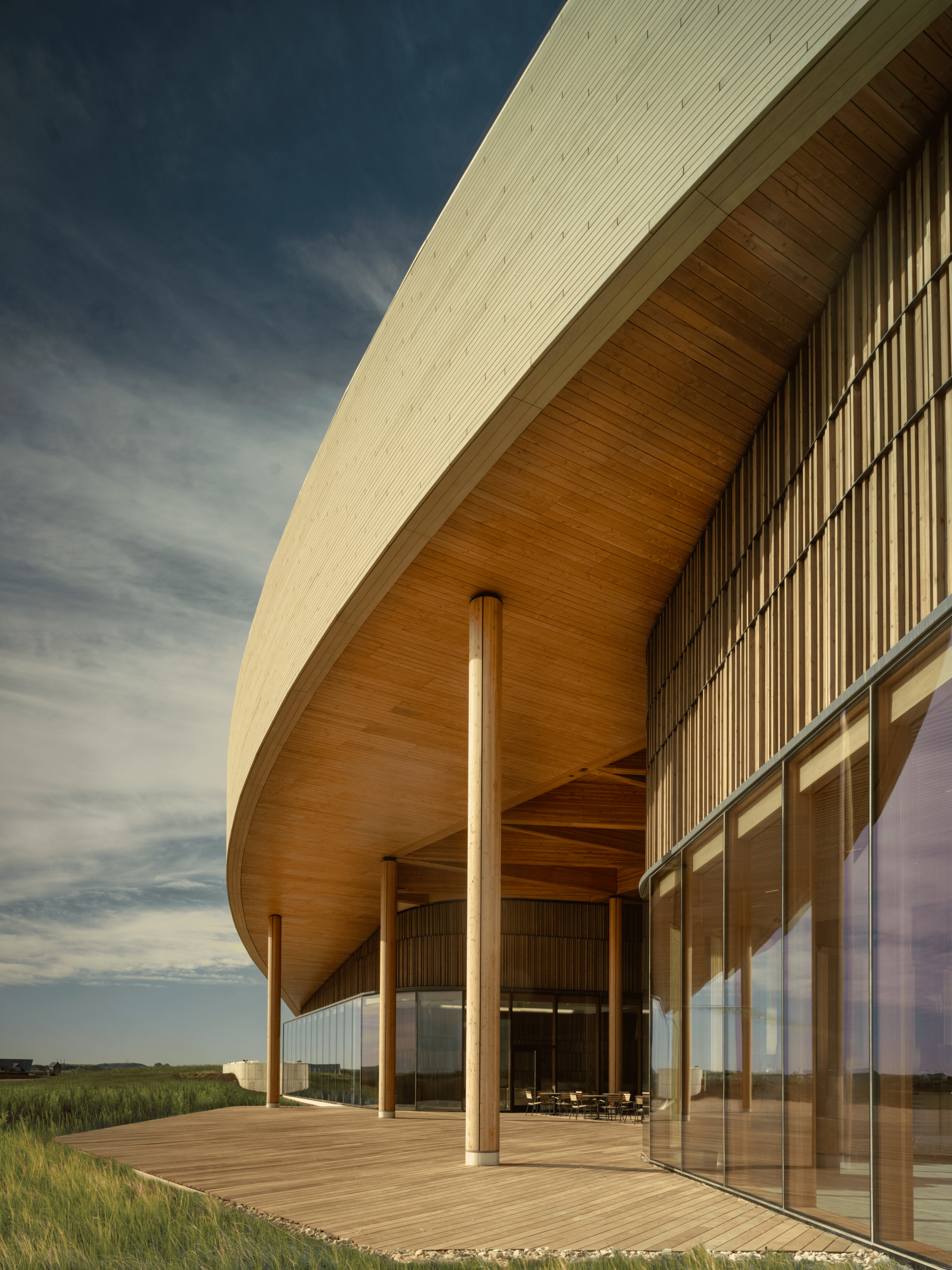 Underside of the curved roof eave with timber soffit, slender wood columns and vertical slat facade beside a glass wall