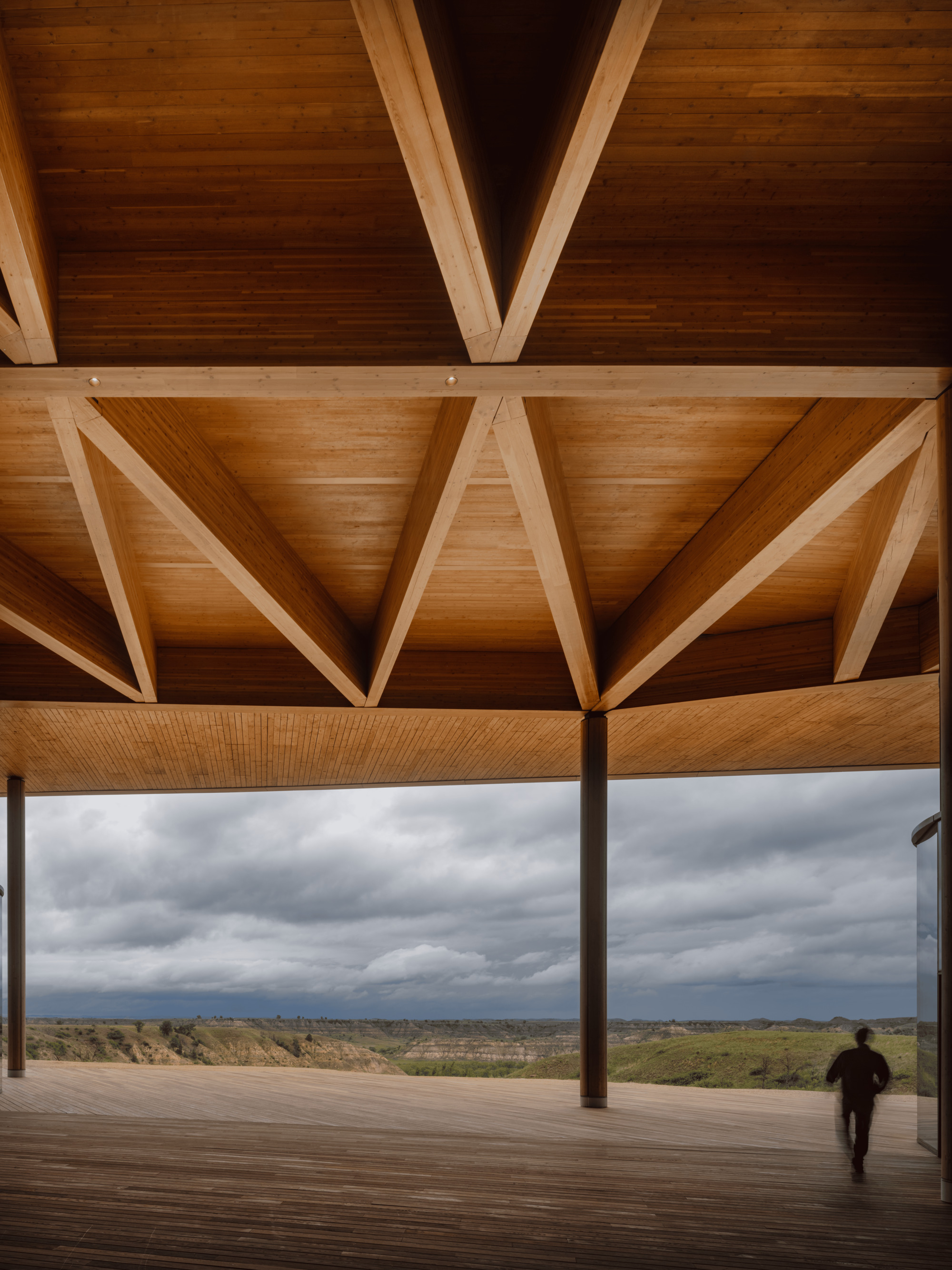 Covered timber deck with exposed glulam beams and a column framing a view of badlands under stormy clouds, a figure walking