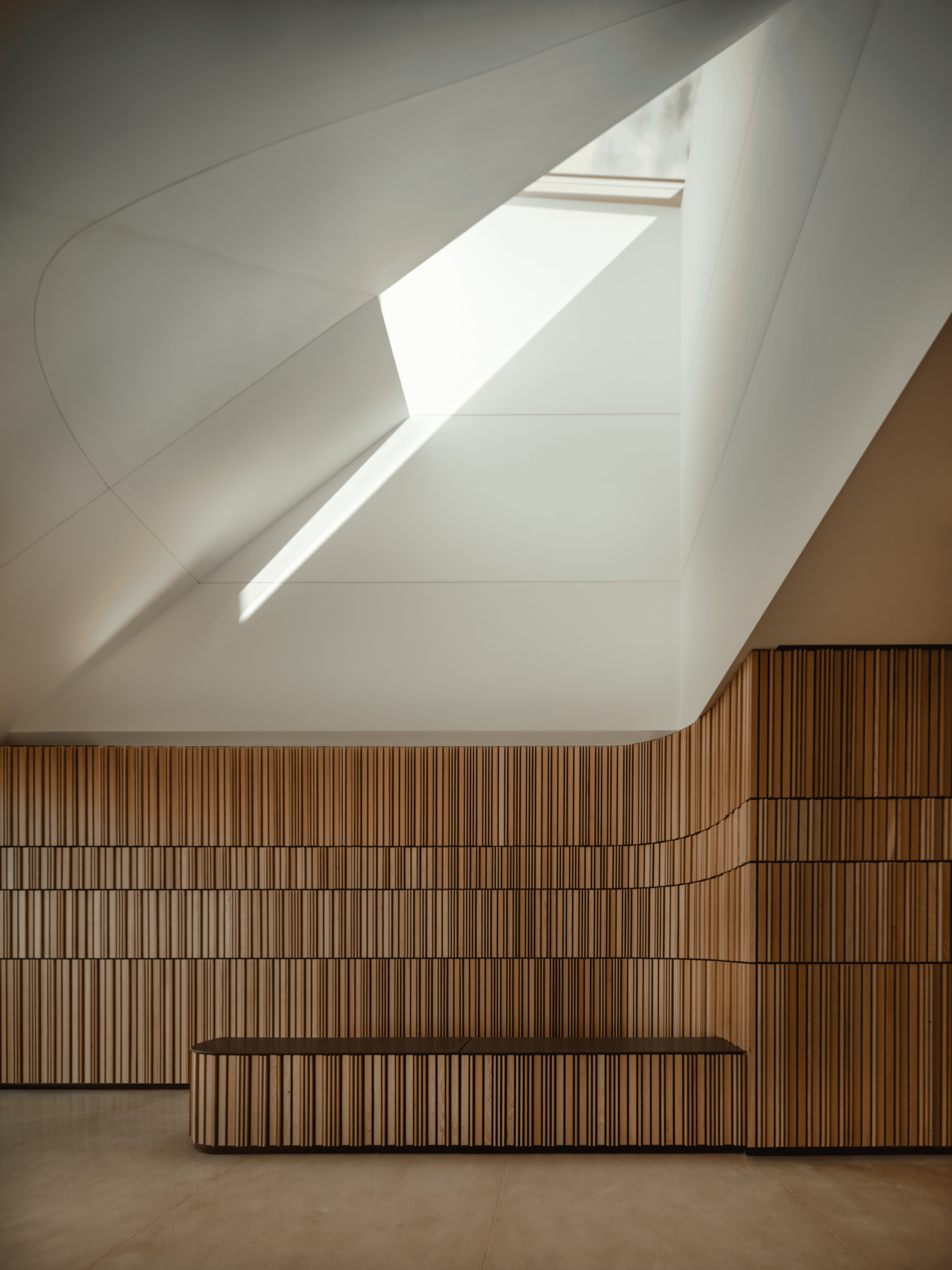 Interior wall of vertical wood slats beneath a white faceted ceiling, with a bench and a beam of sunlight from a skylight