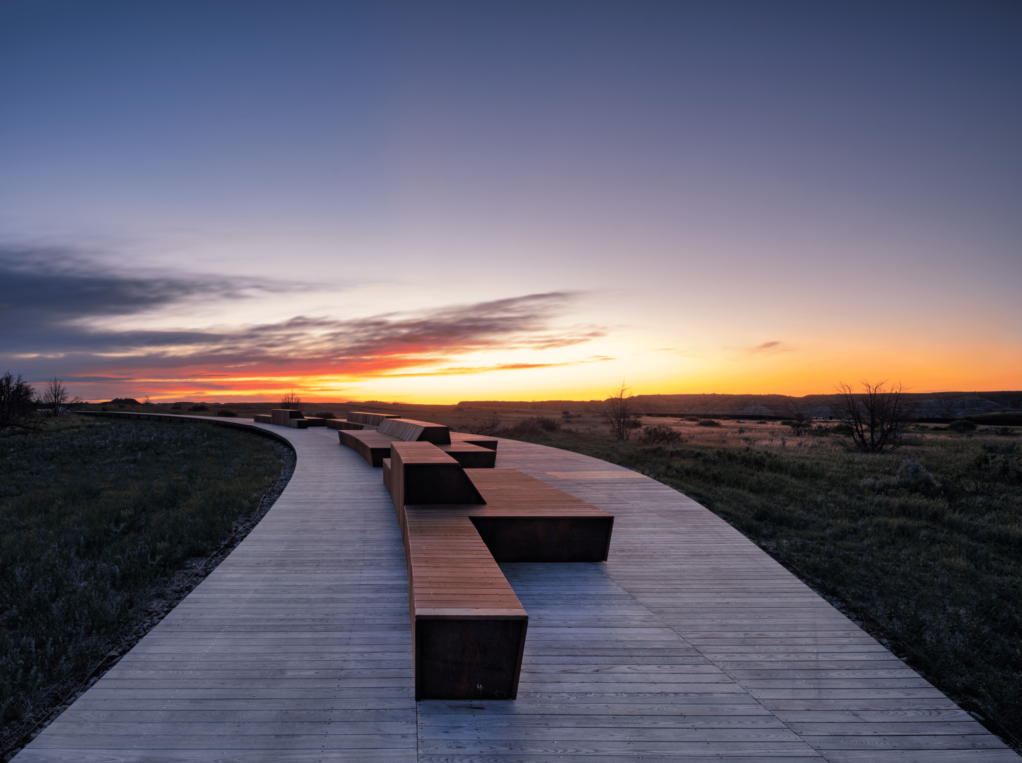 Curving timber boardwalk with angular wooden benches at sunset, orange light low over the prairie horizon