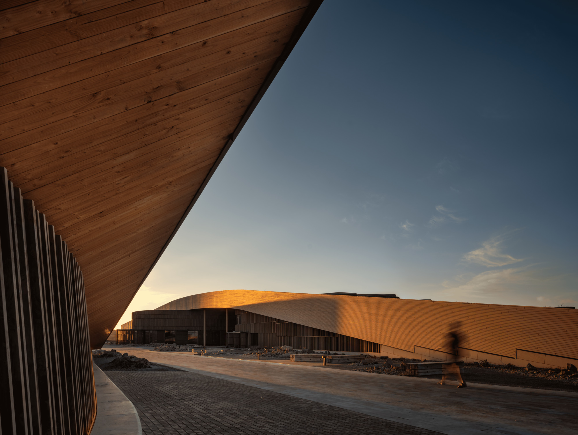 Timber-clad soffit and slatted wall frame a curved wooden roof, with a blurred figure walking past at dusk