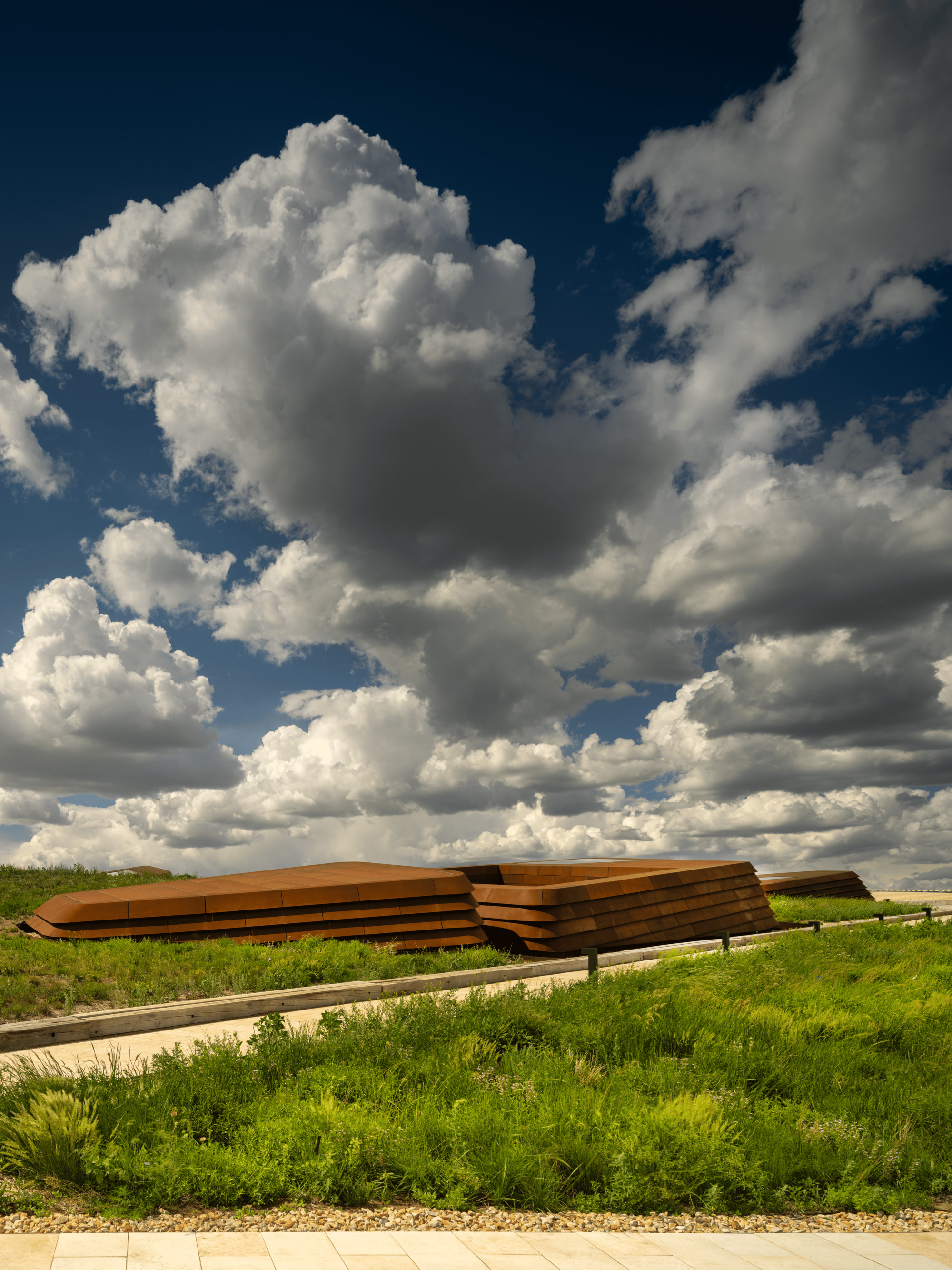 Stacked weathering steel layers forming a low sculptural volume behind green grasses under towering white clouds