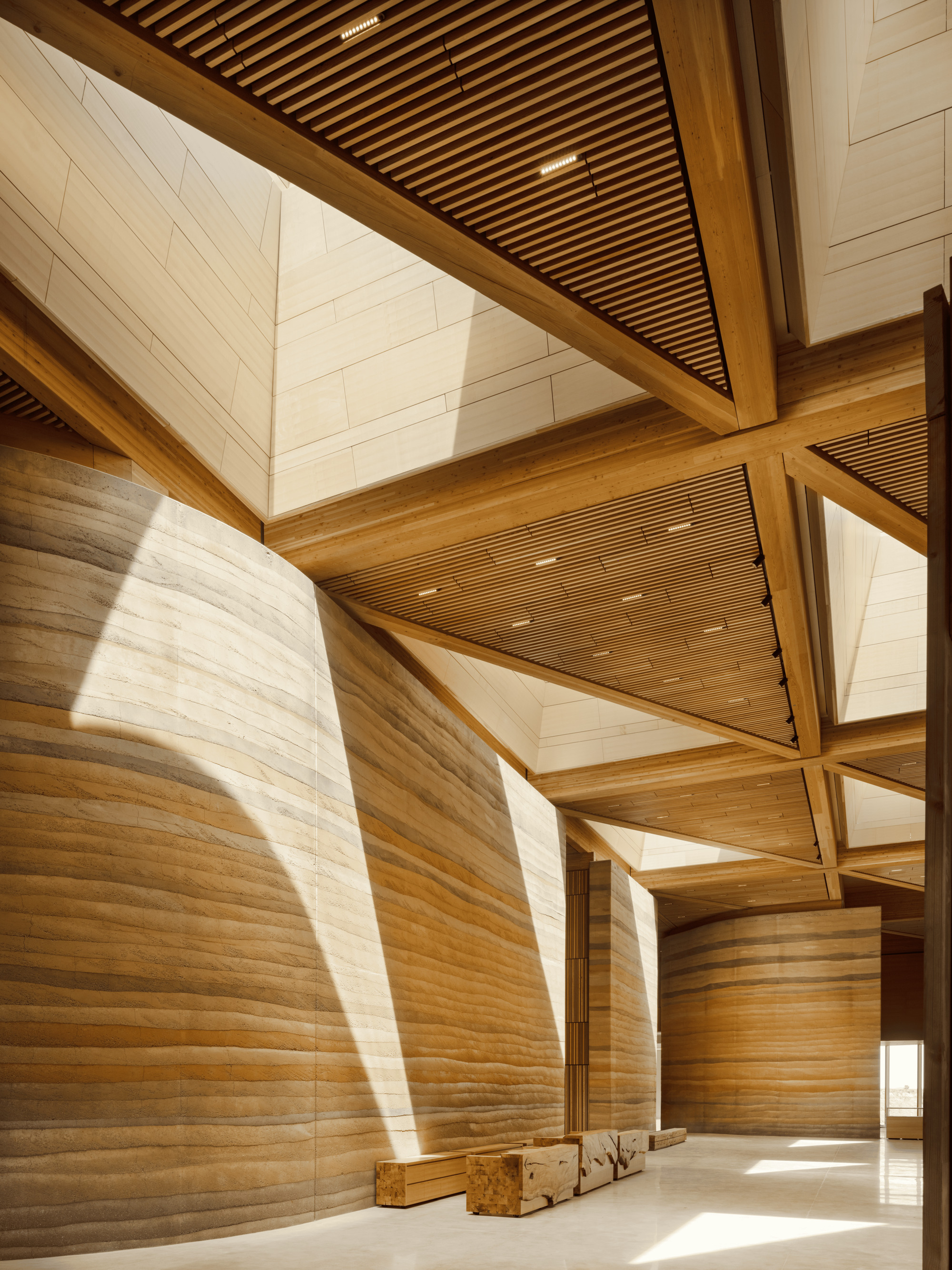Interior hall with curved rammed earth walls, glulam timber beams and slatted ceiling panels, with sunlight angling across the stone floor