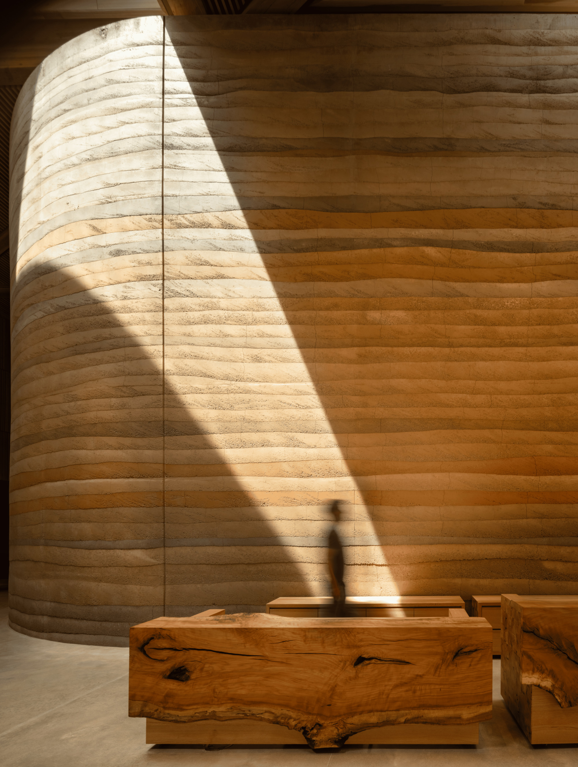 Layered rammed earth curved wall cut by a diagonal beam of sunlight, with a blurred visitor behind rough-edged timber benches