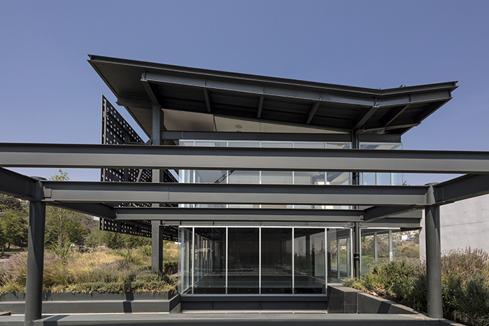 Steel beams frame the glazed pavilion beneath a butterfly-style roof, with grasses in planters under a clear blue sky