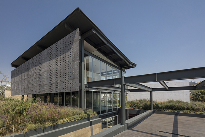 Corner of the pavilion showing the perforated screen, glass walls and steel beams reaching over a timber deck beside planted beds