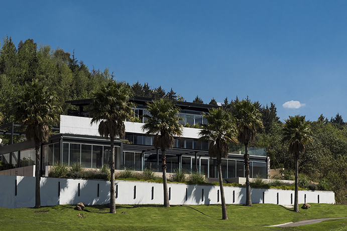 Hillside house with long glazed terraces above a white slotted wall, framed by palm trees and a forested slope