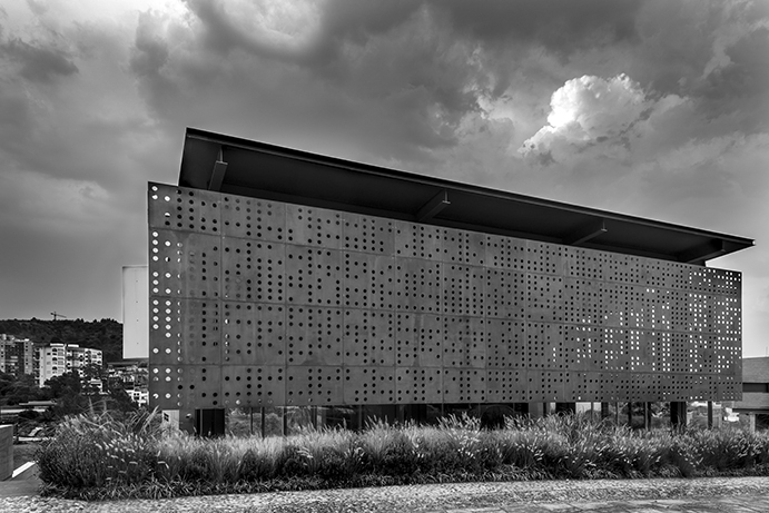 Black and white view of the perforated box facade under a deep cloudy sky, with grasses in the foreground