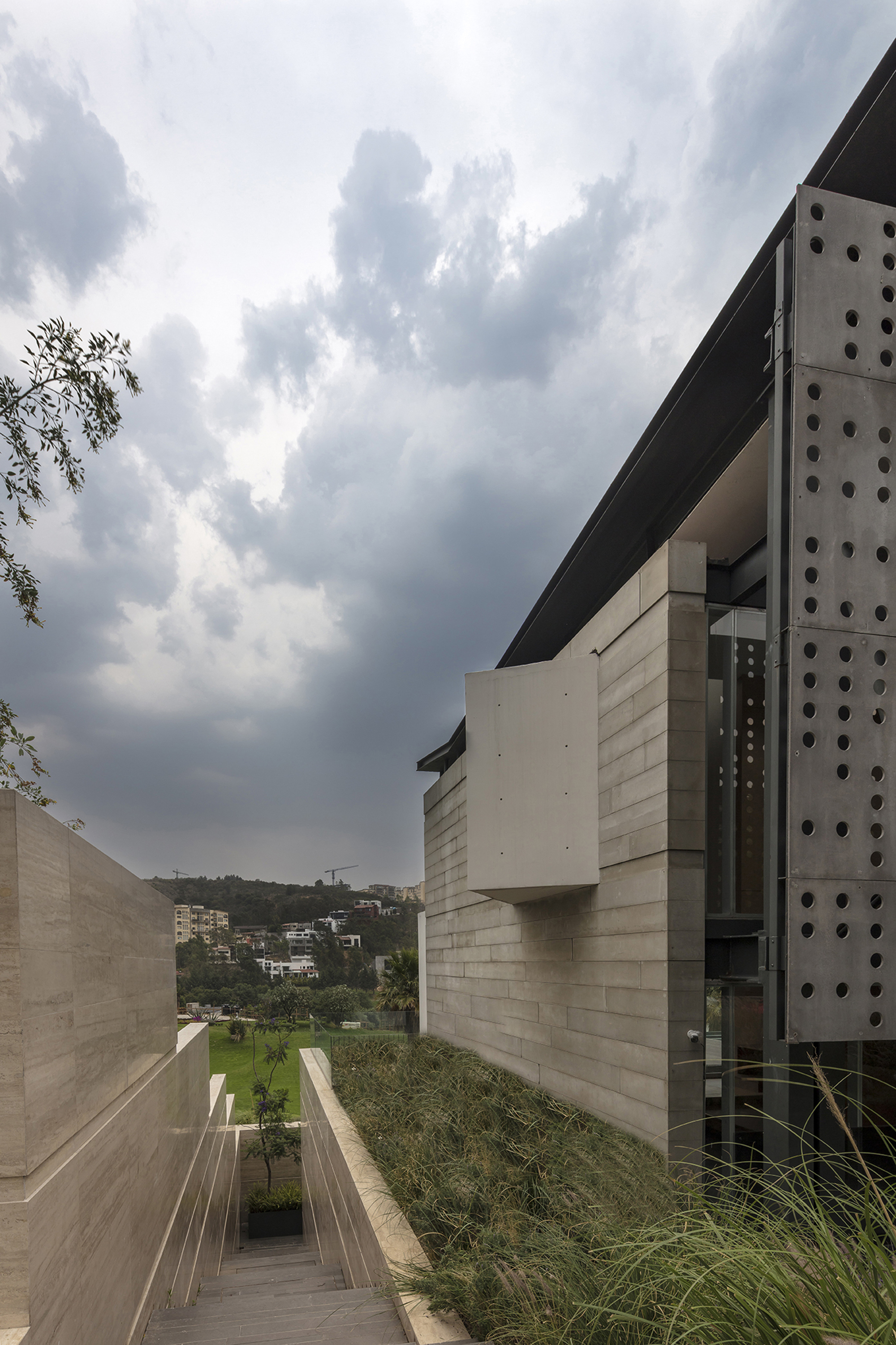 Narrow gap between a travertine-clad wall and a perforated steel screen, with grasses in the foreground under heavy storm clouds
