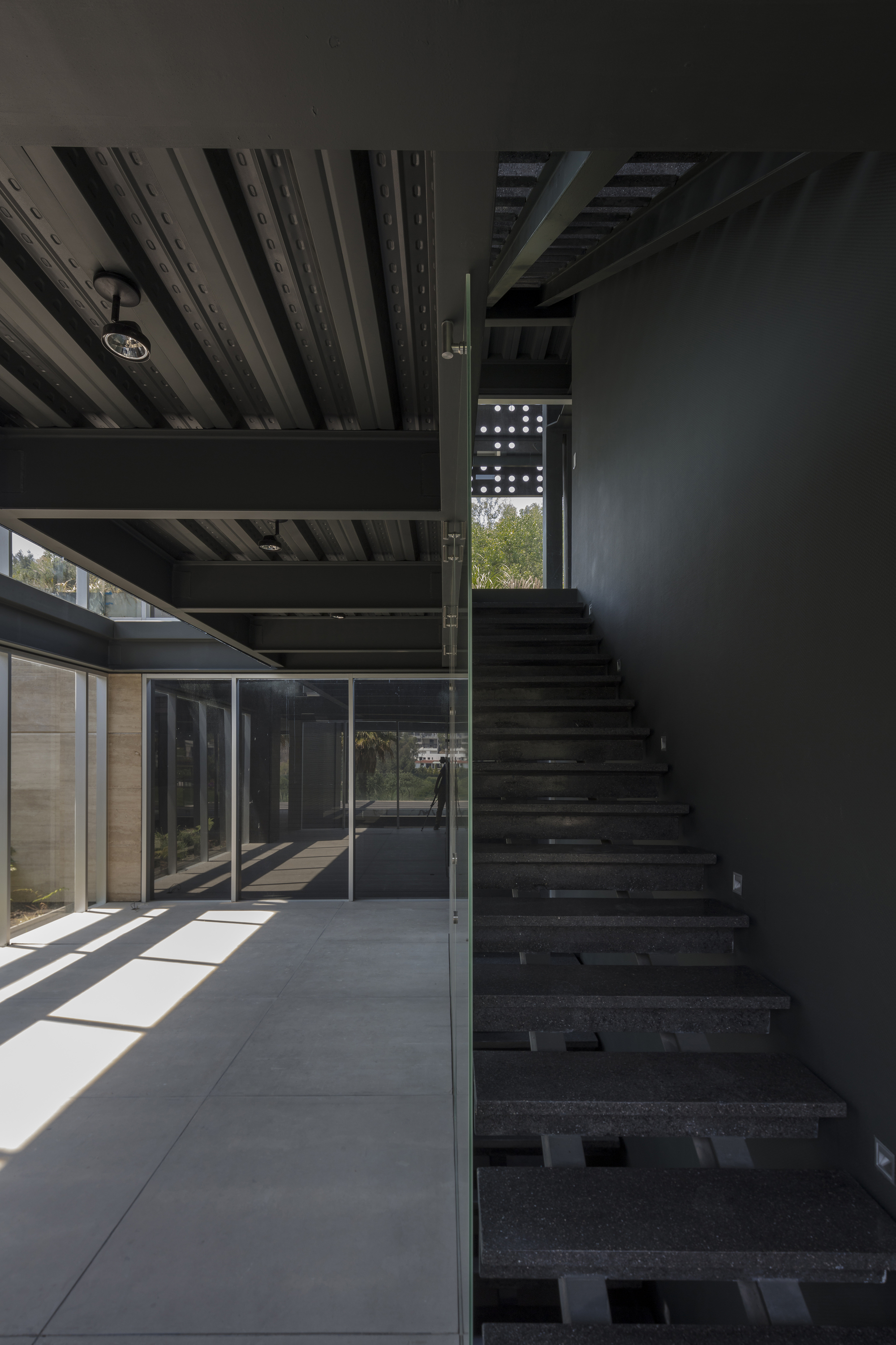 Open steel stair with dark stone treads beside a glass balustrade, with sunlight falling across the concrete floor
