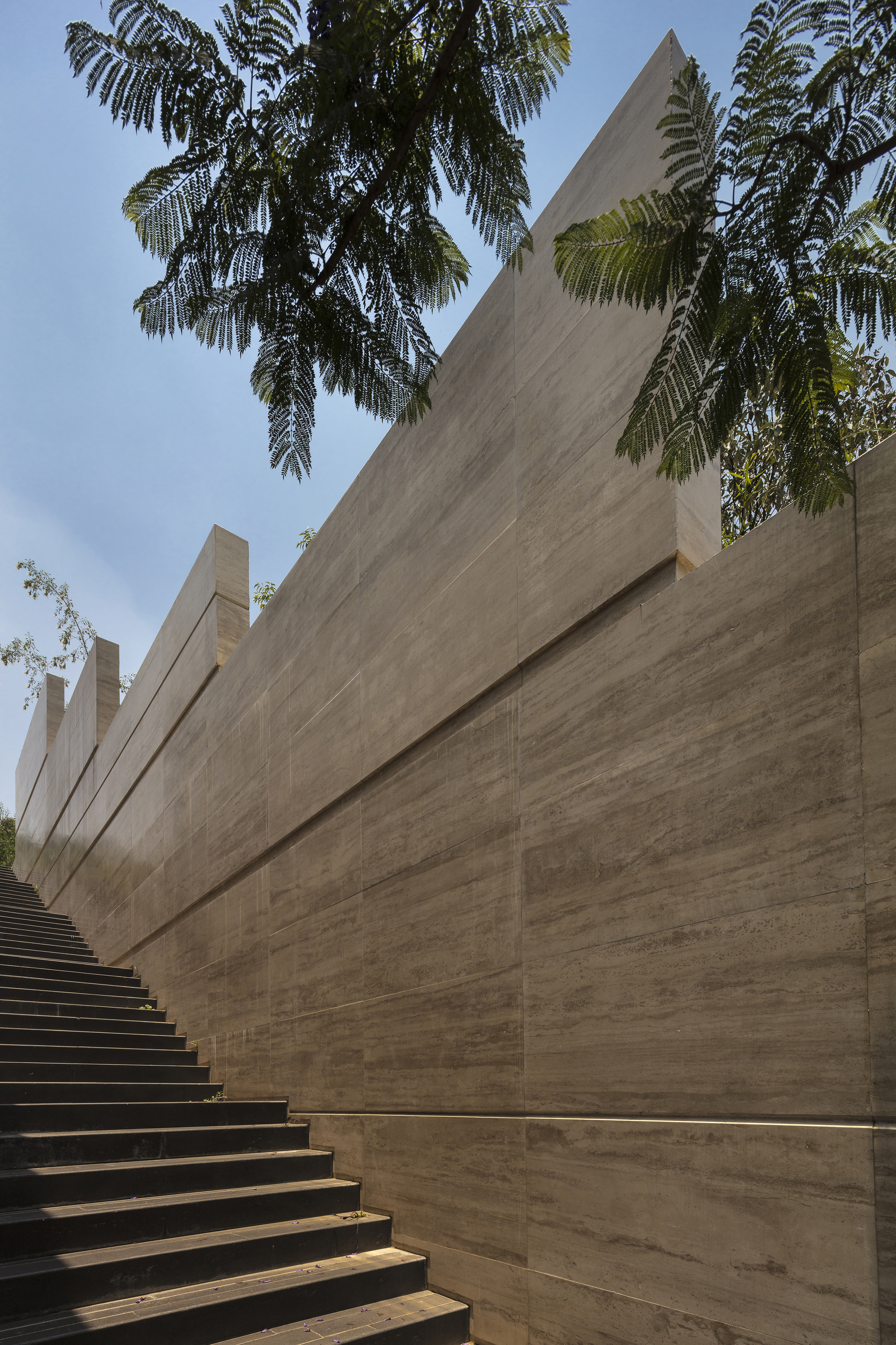 Stepped travertine wall rising alongside a dark stair, framed by fern-like tree branches against a blue sky