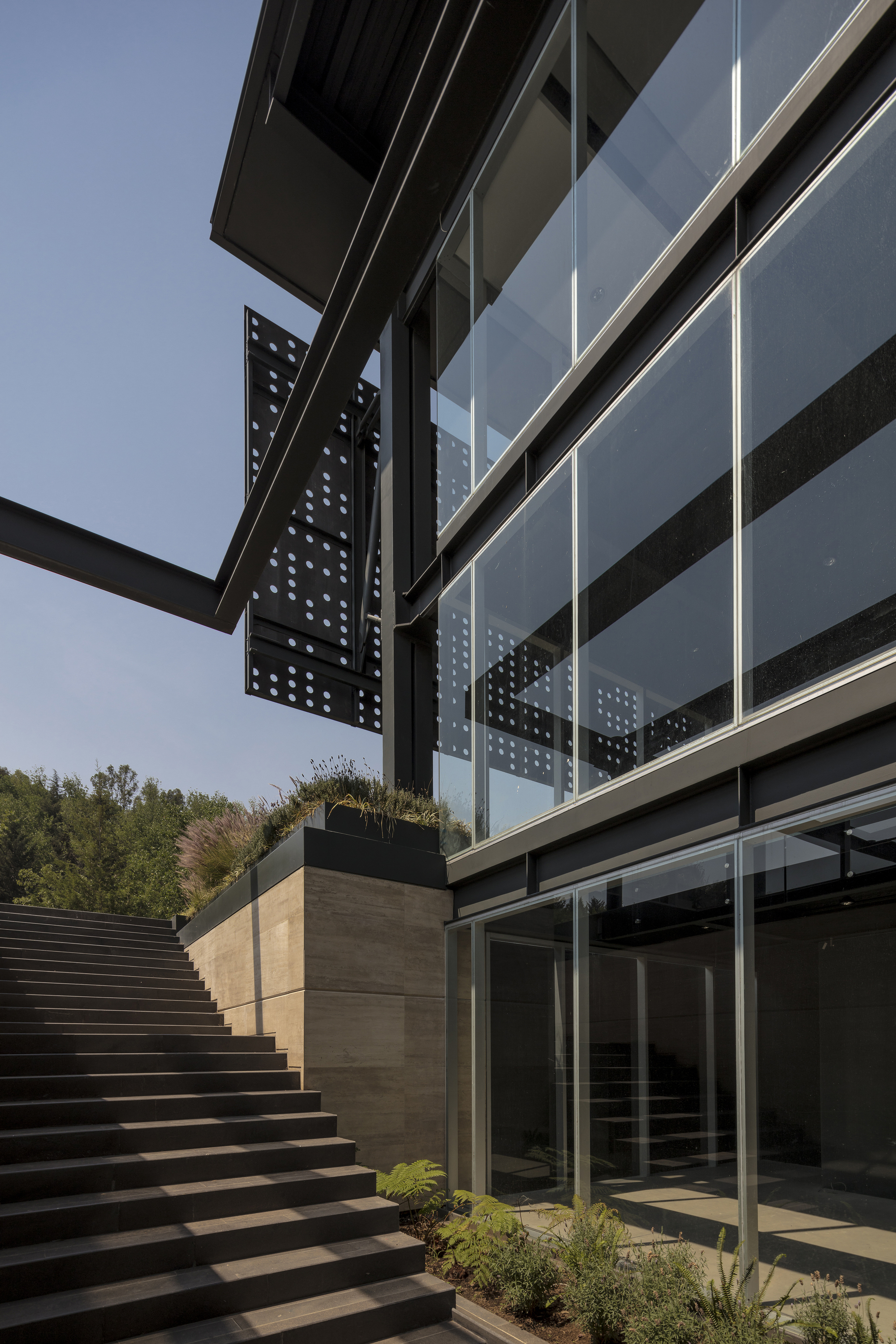 Glazed facade with a perforated steel panel above a planted travertine terrace and wide stair under clear sky