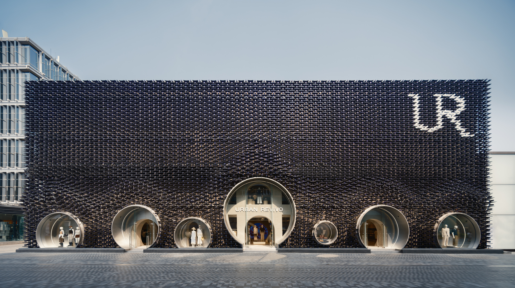 Frontal view of the black studded facade with round porthole display windows and an illuminated letter logo under a pale sky