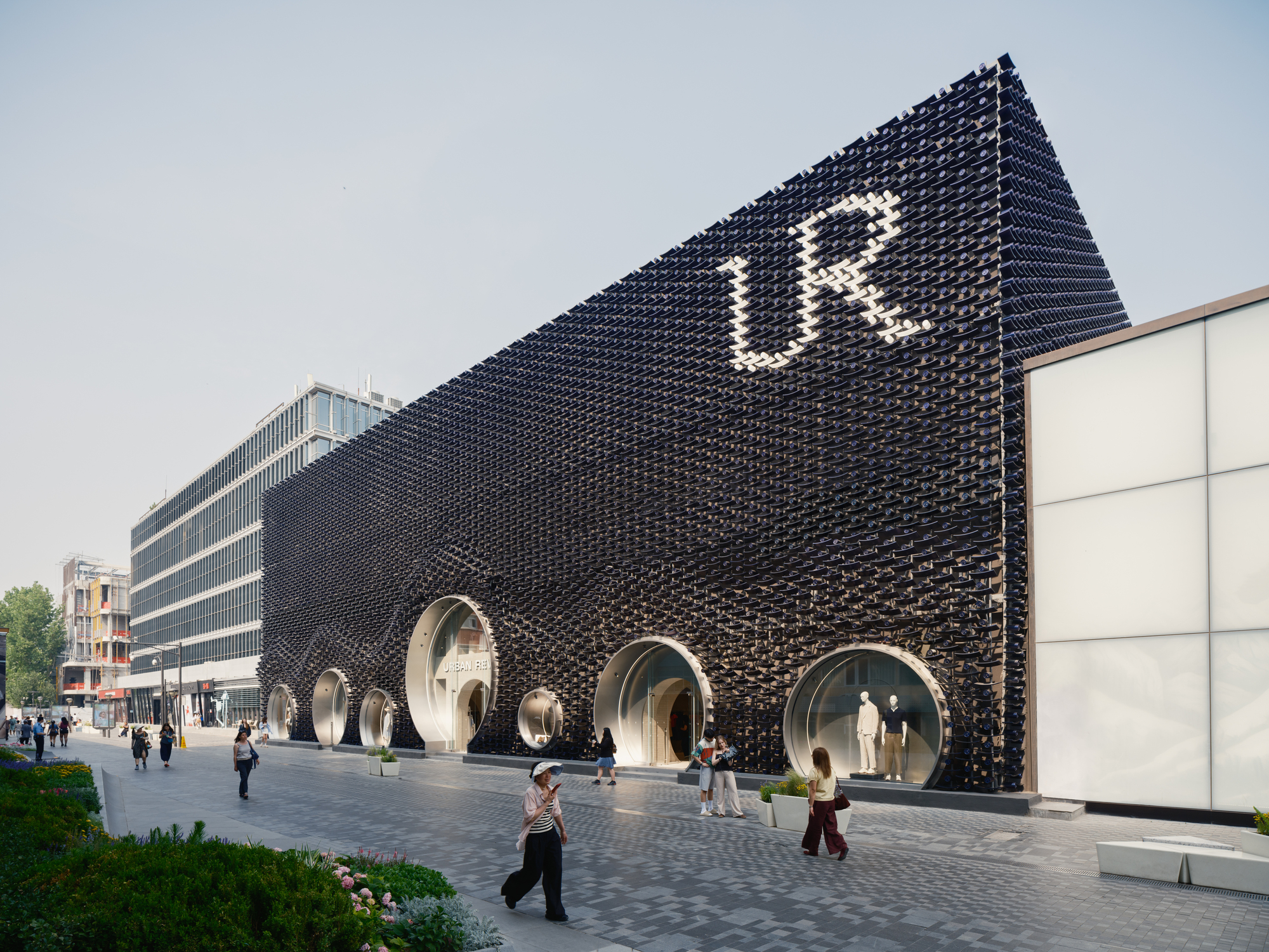 Angled view of the dark textured facade and circular shopfront openings, with pedestrians crossing the paved street in hazy daylight