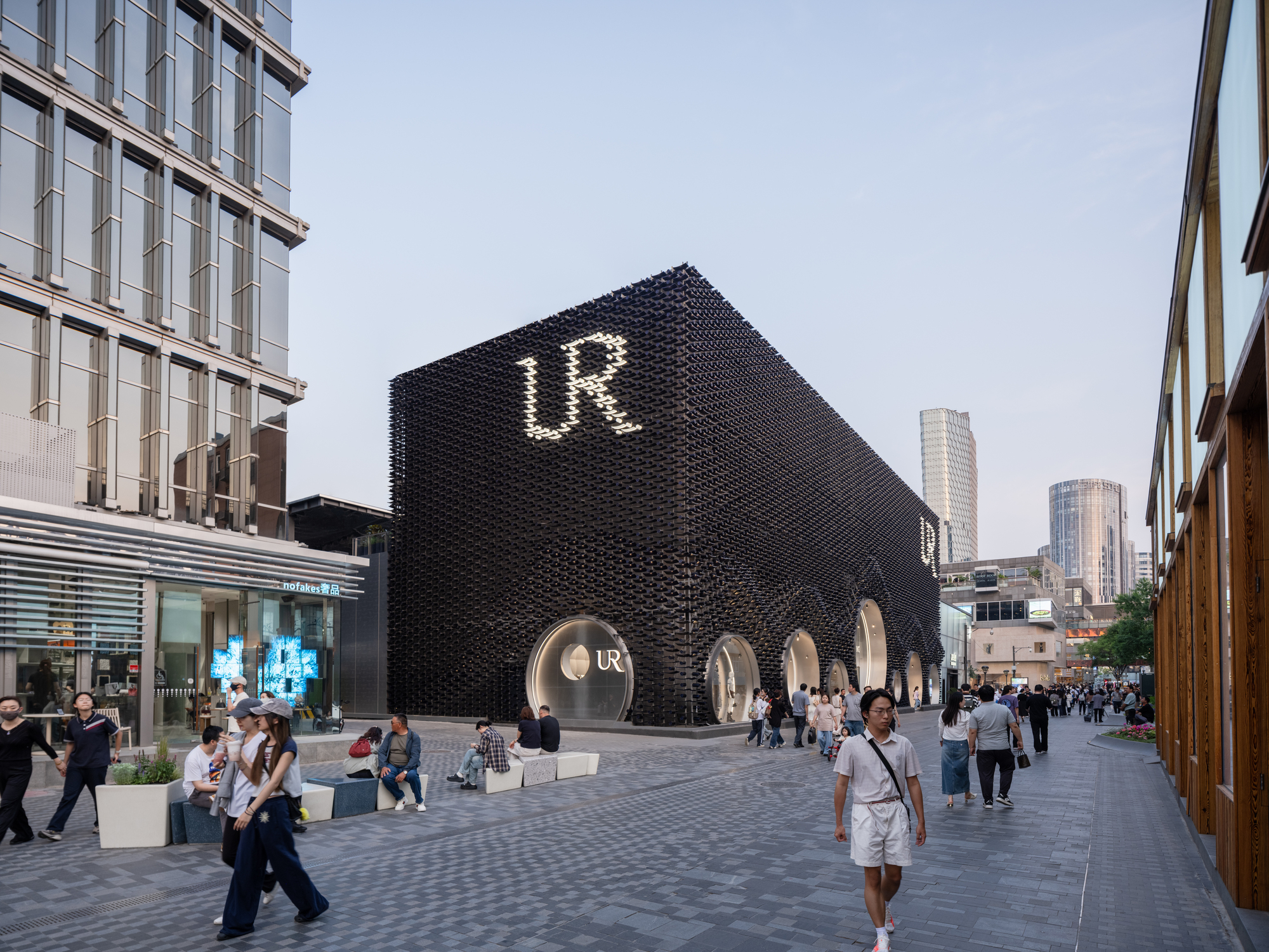 Corner view of the black box facade with glowing letter sign and round windows, shoppers walking the paved plaza at dusk