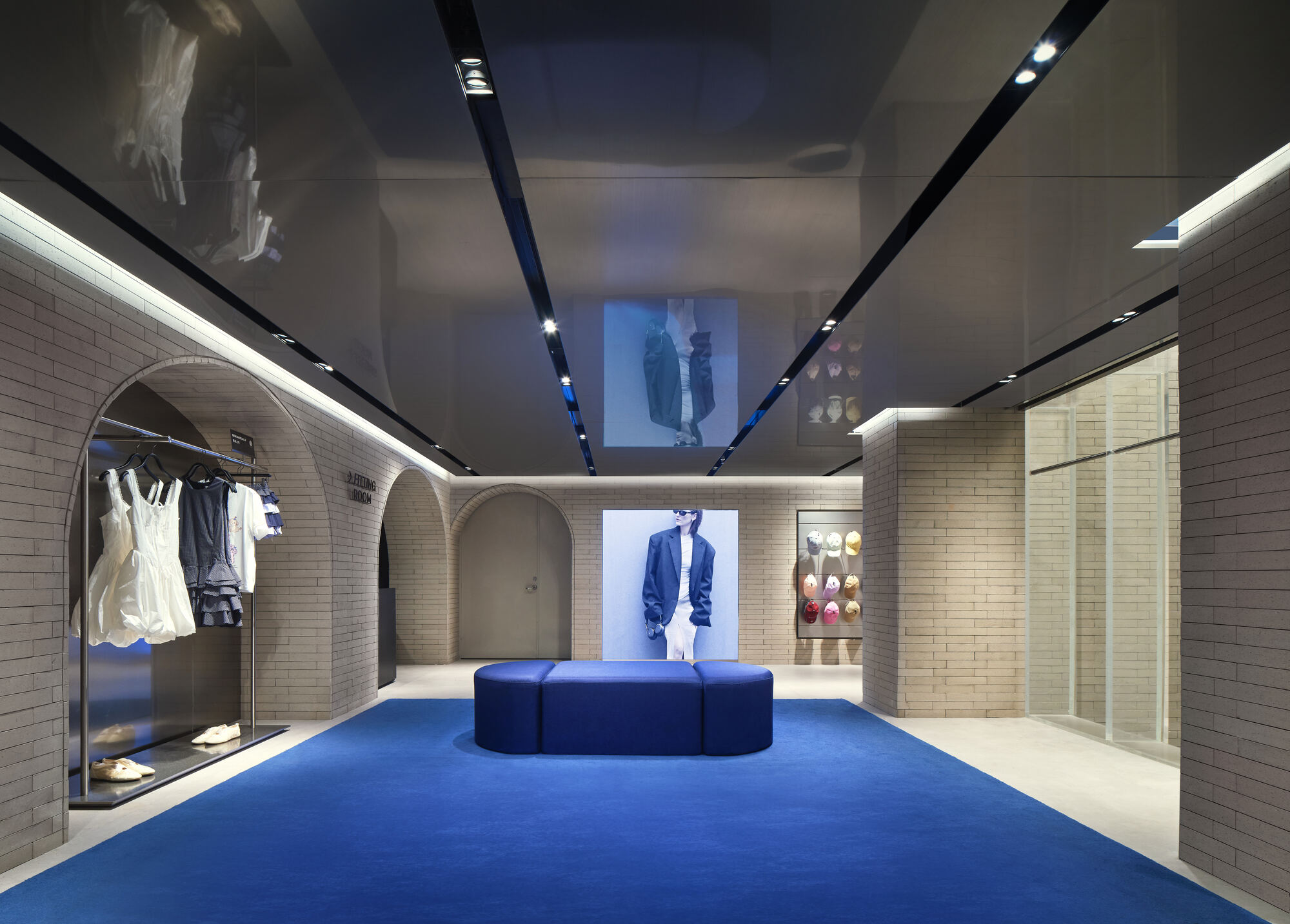 Gallery with arched brick alcoves, a blue carpet and bench, and a mirrored ceiling with linear light slots reflecting garments