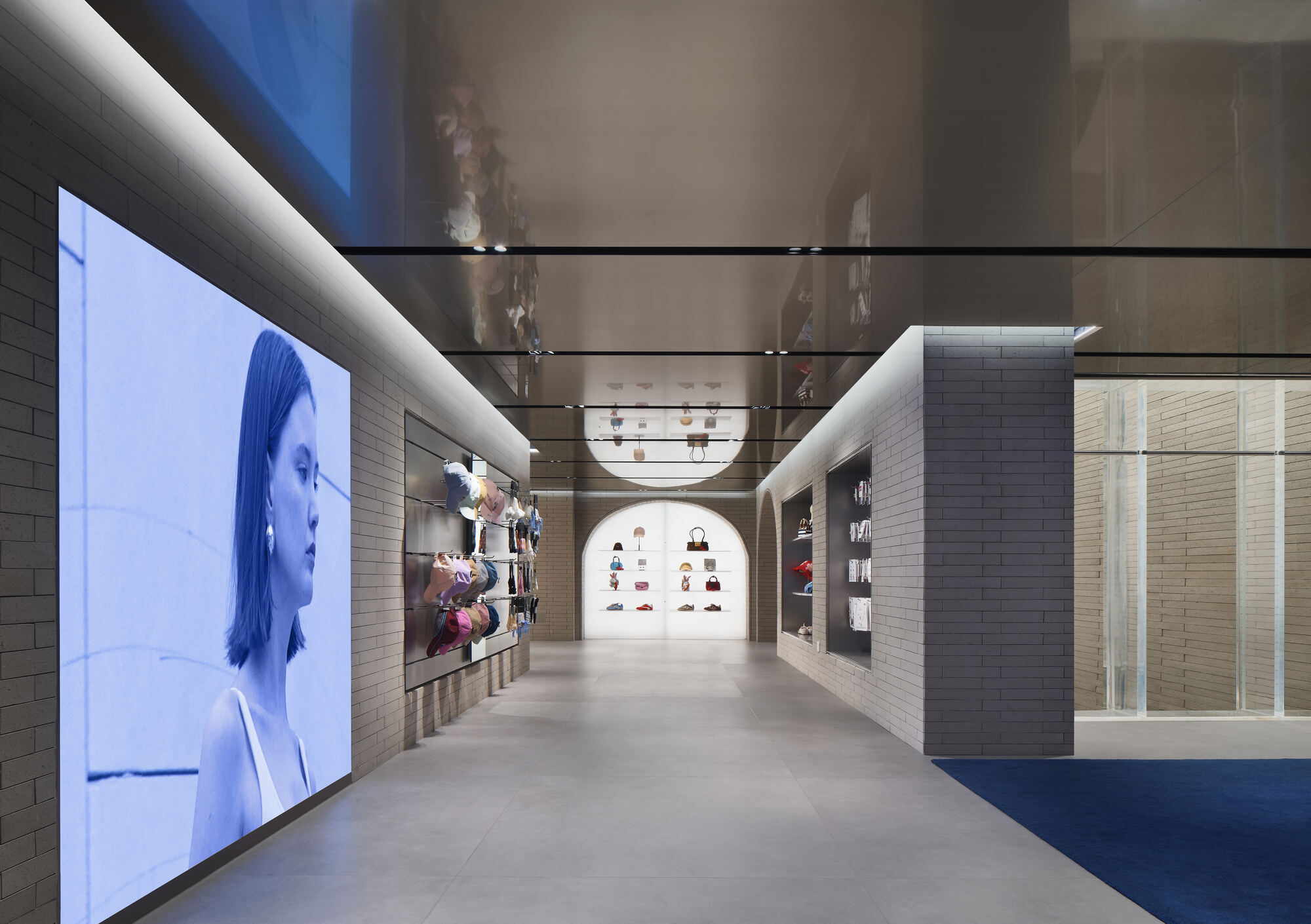 Brick-walled corridor with a large screen showing a woman in profile, a mirrored ceiling and a lit arched niche of handbags at the end