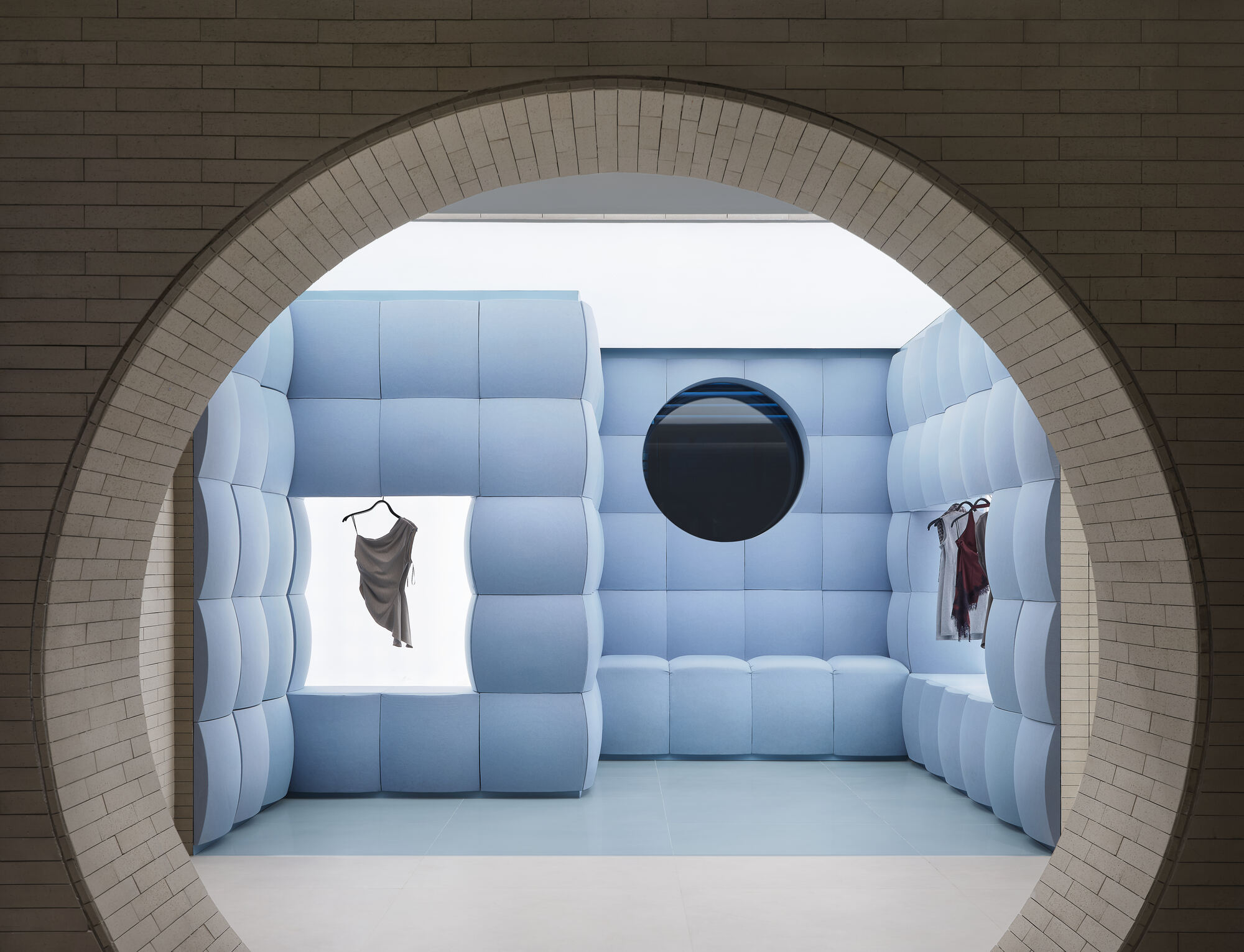 Circular brick opening framing a pale blue quilted-panel alcove with a round mirror and garments hung in lit recesses
