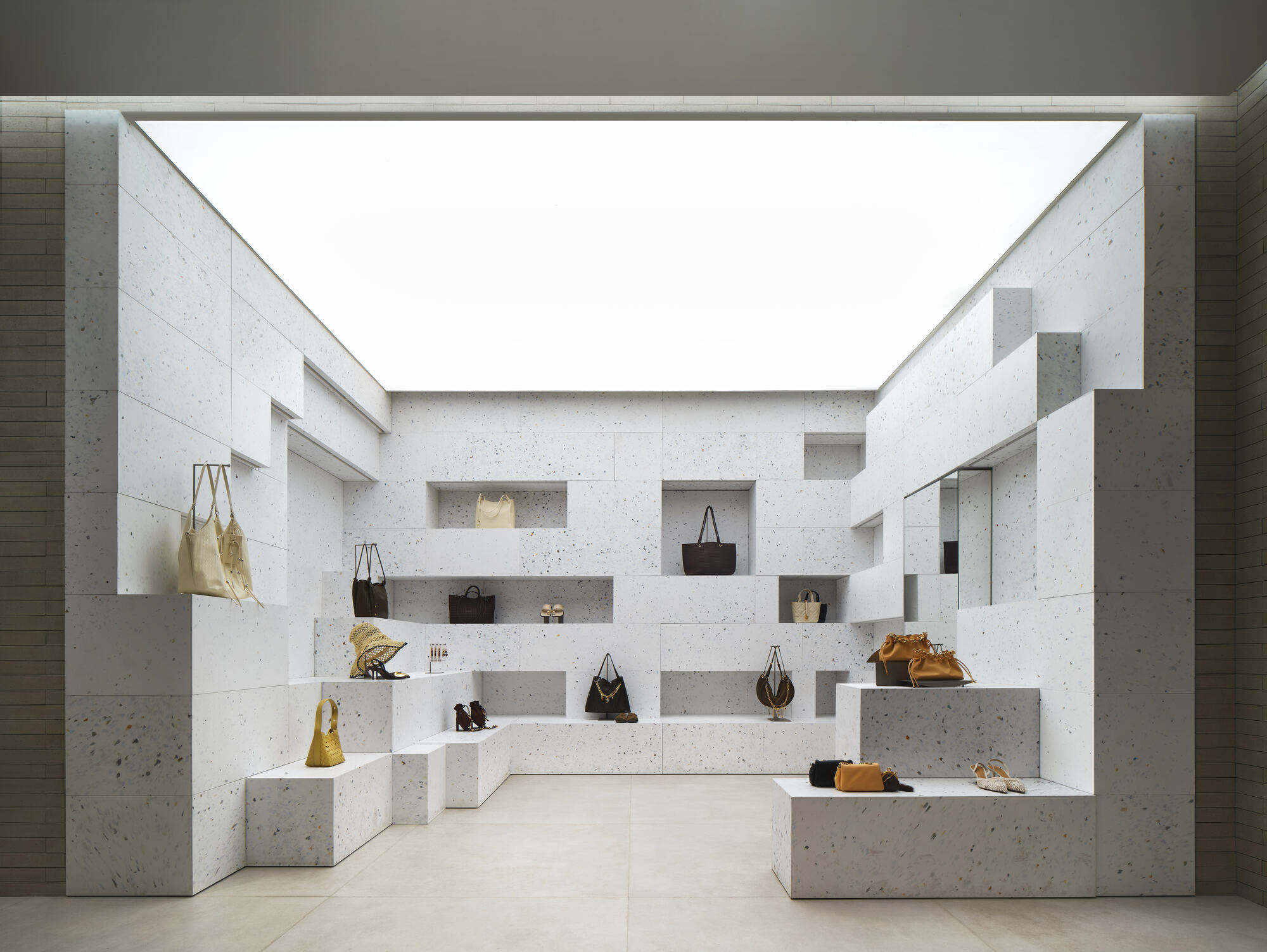 Display niche of speckled white stone blocks holding handbags and shoes beneath a glowing luminous ceiling panel