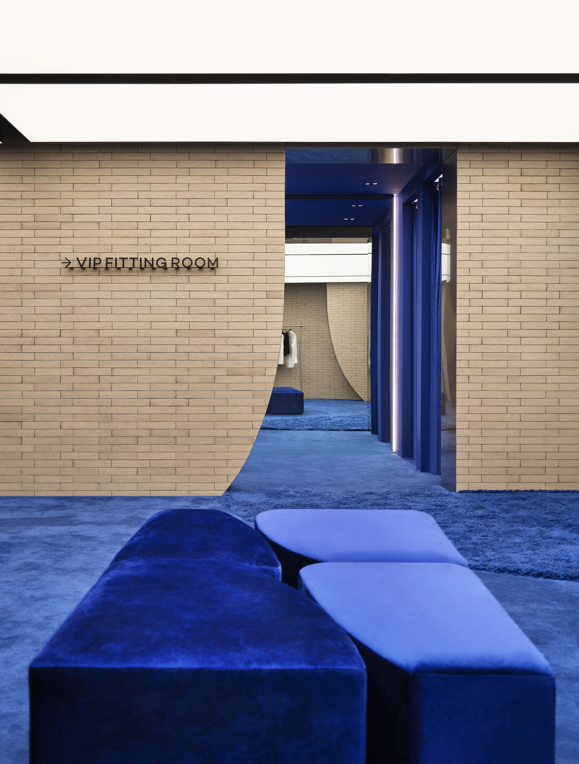 Brick wall with a curved cut leading to a VIP fitting room, with cobalt velvet ottomans on blue carpet in the foreground