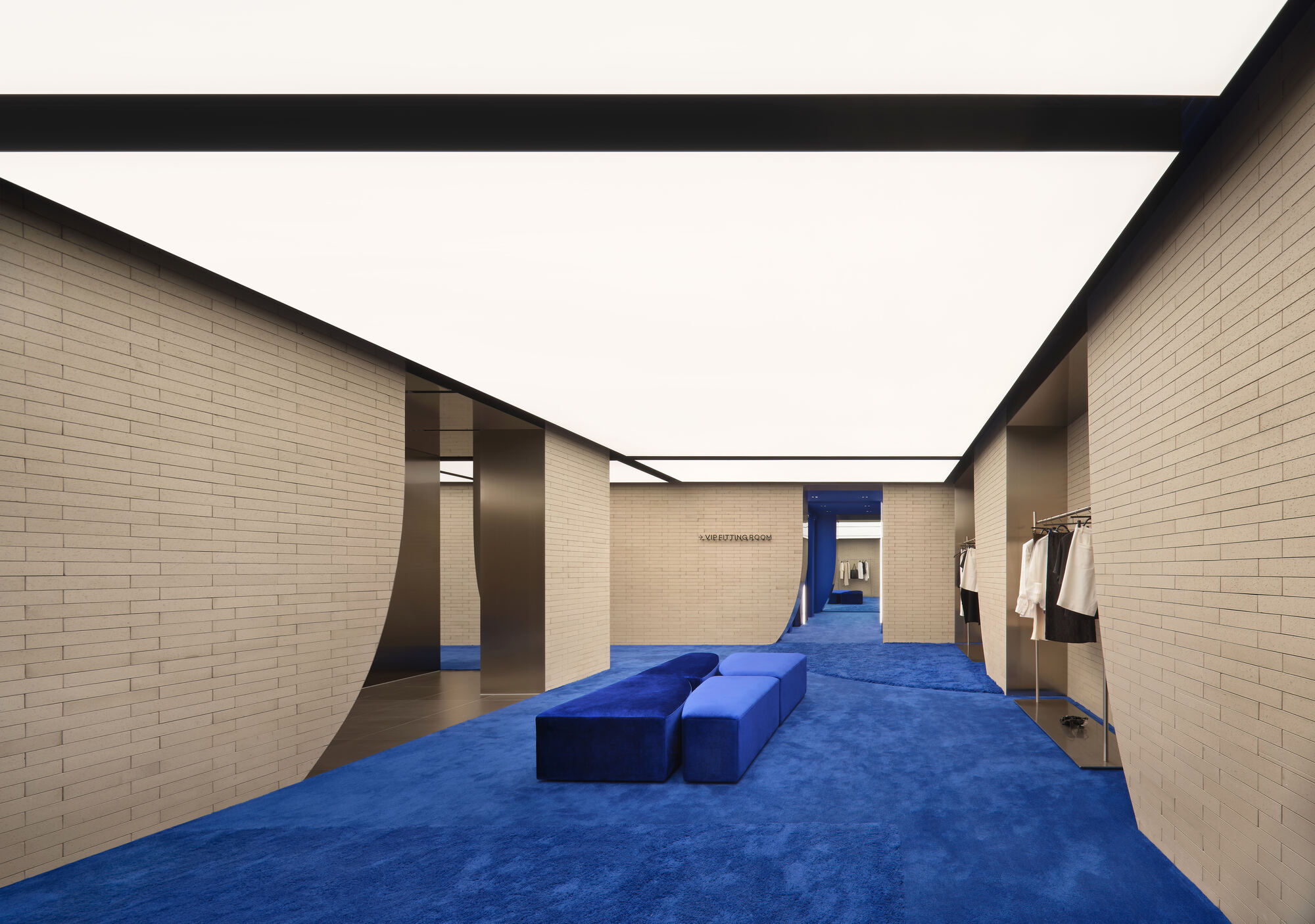 Corridor of pale brick walls with curved cut-outs, cobalt carpet and two velvet benches under a long luminous ceiling