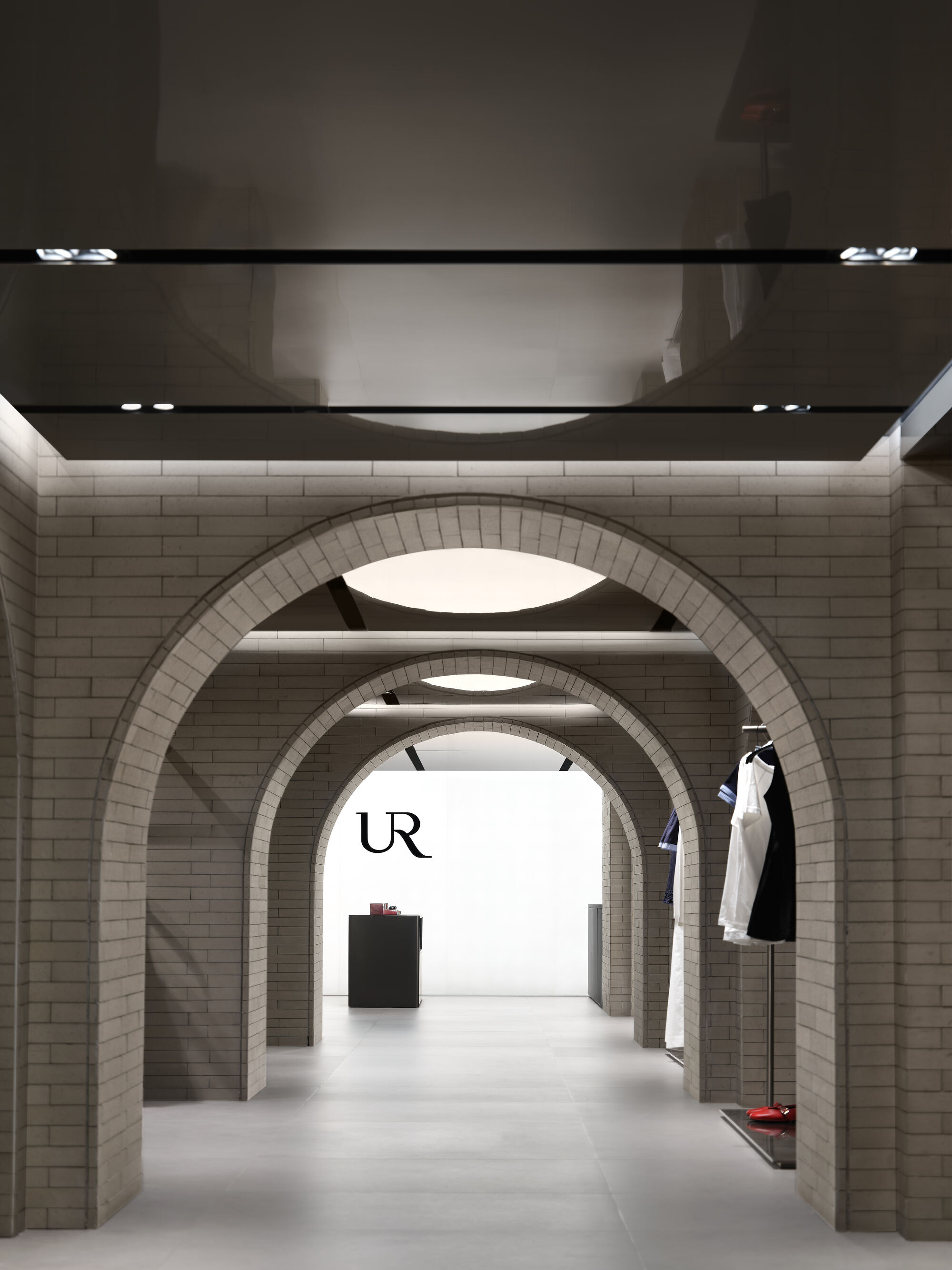 Sequence of three brick arches receding toward a lit logo wall, with a mirrored ceiling above and a garment rack at right