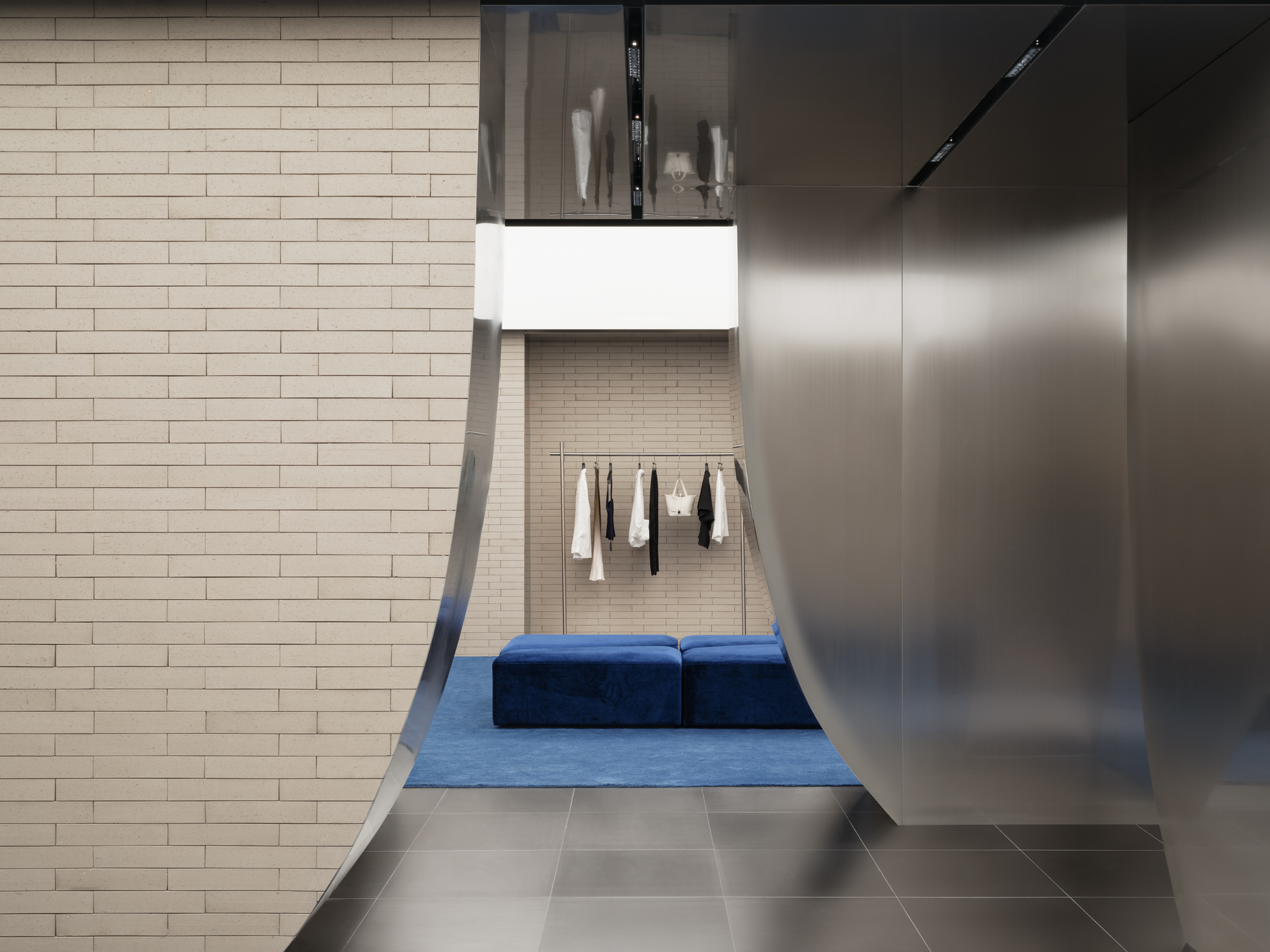 Curved brushed steel panels beside a brick wall, framing a blue velvet bench and a rack of clothes