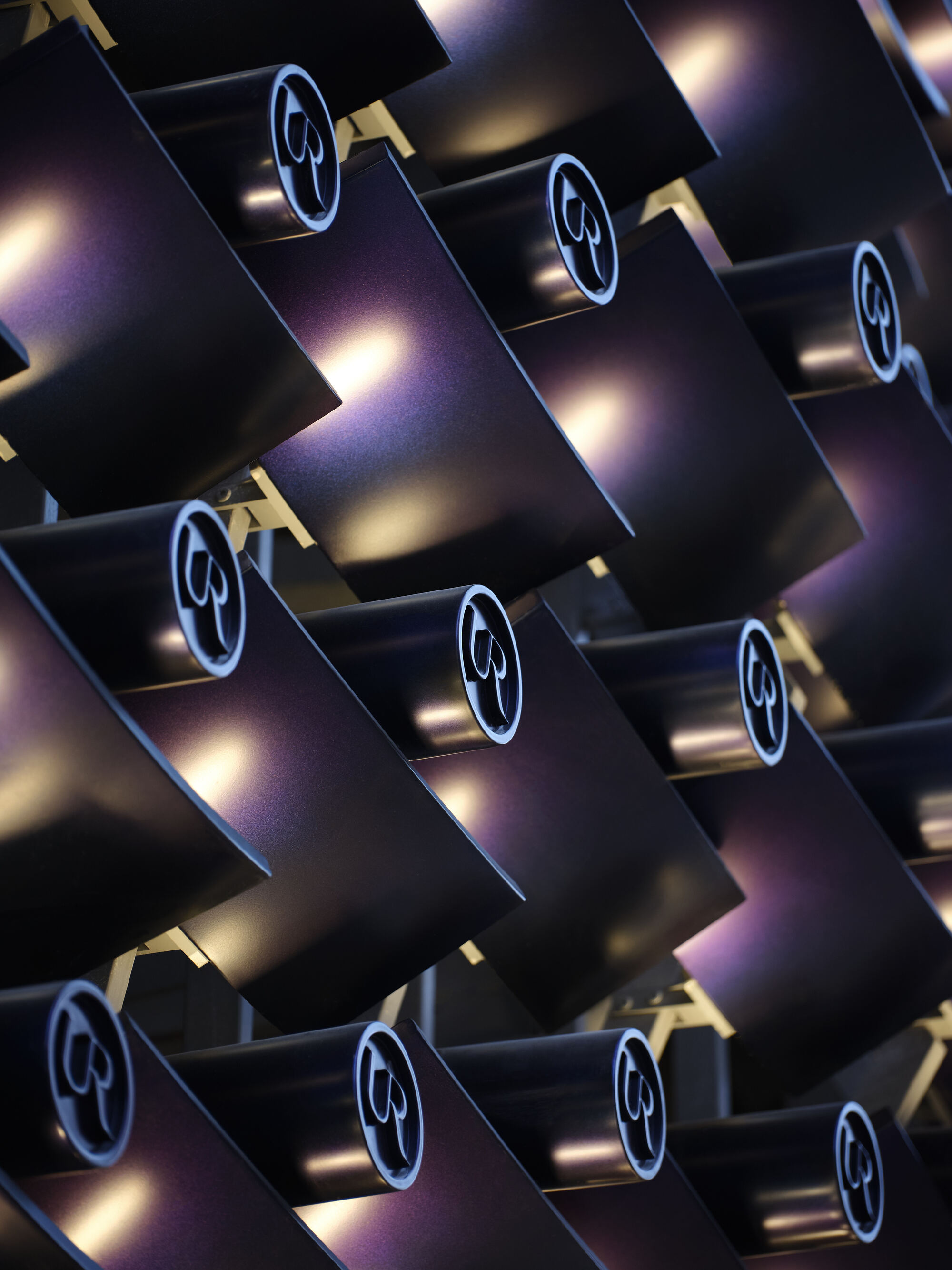 Close-up of overlapping curved purple-black metal panels, each fitted with a round logo disc, catching warm light