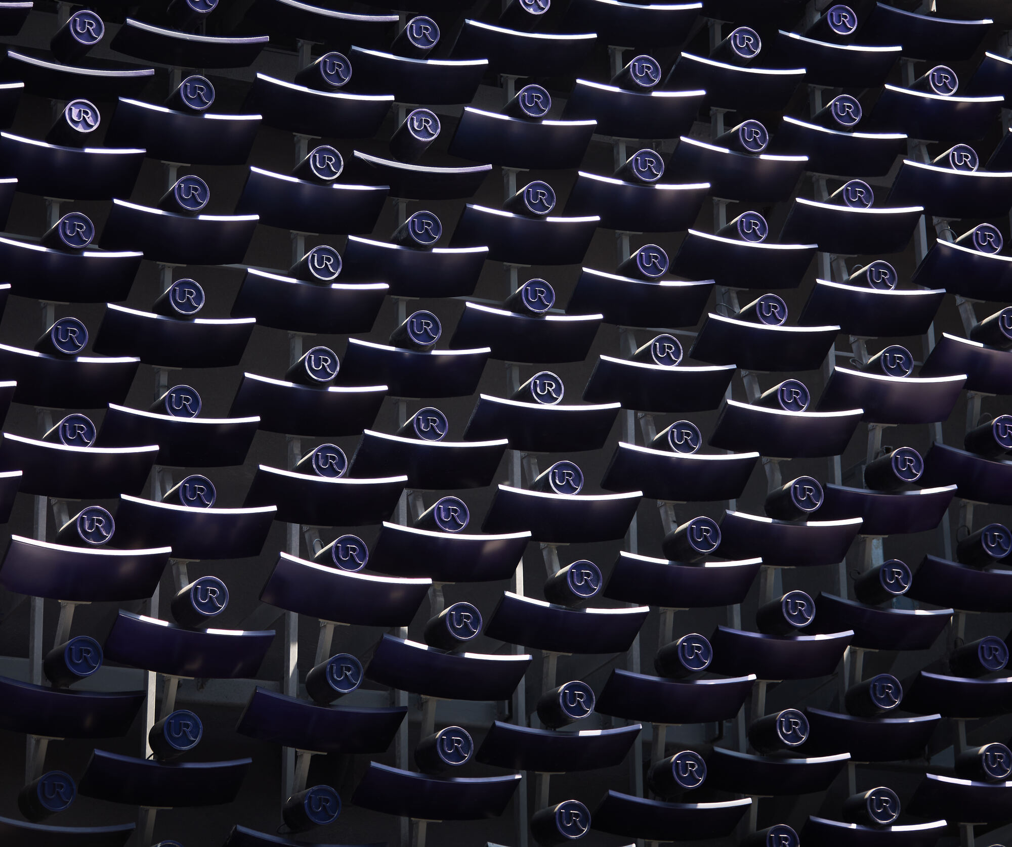 Dense grid of dark curved blades with round logo discs, their upper edges lit by a thin line of light