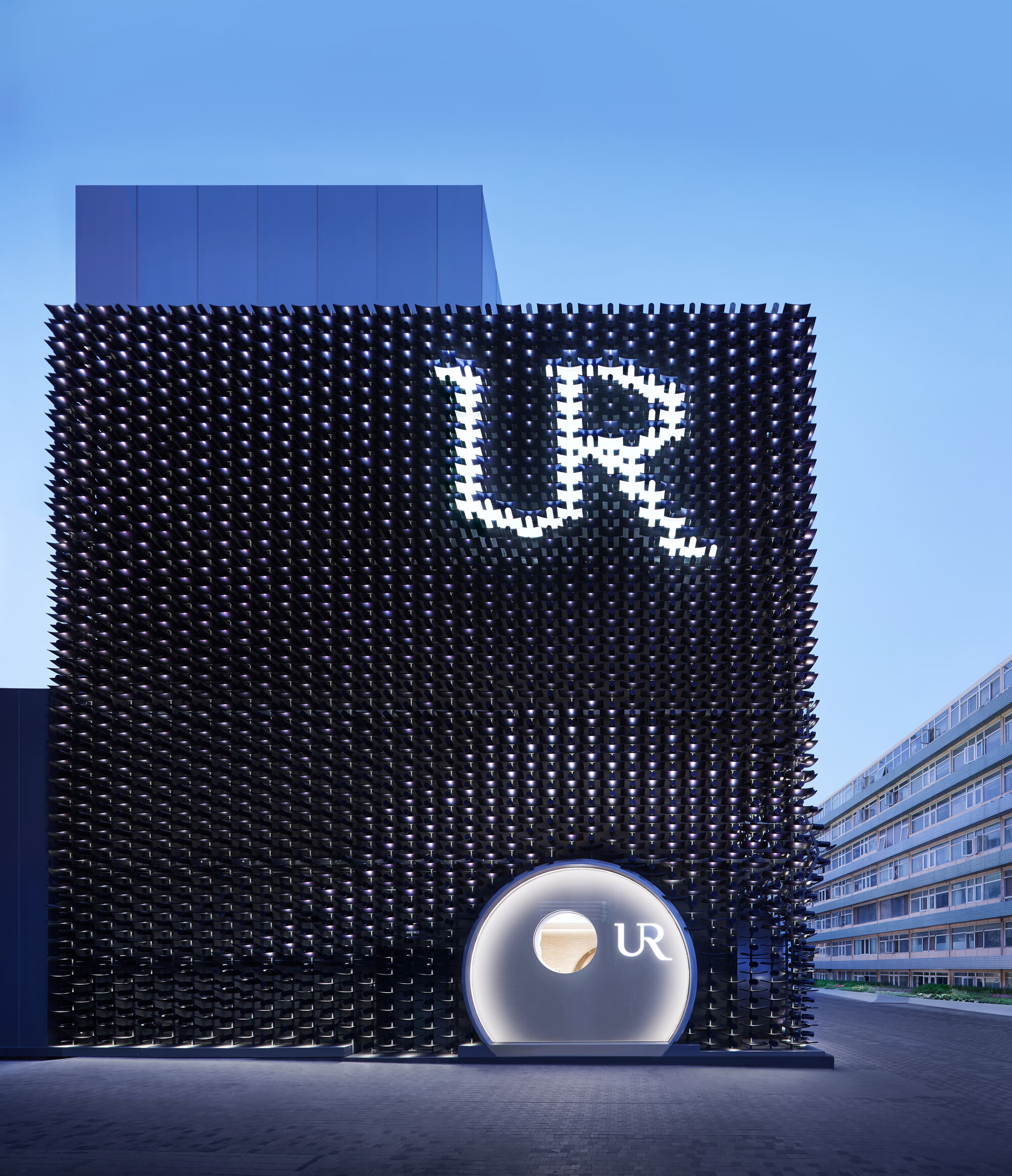 Front face of a cube clad in black protruding pegs, with a lit letter sign and round entrance at dusk