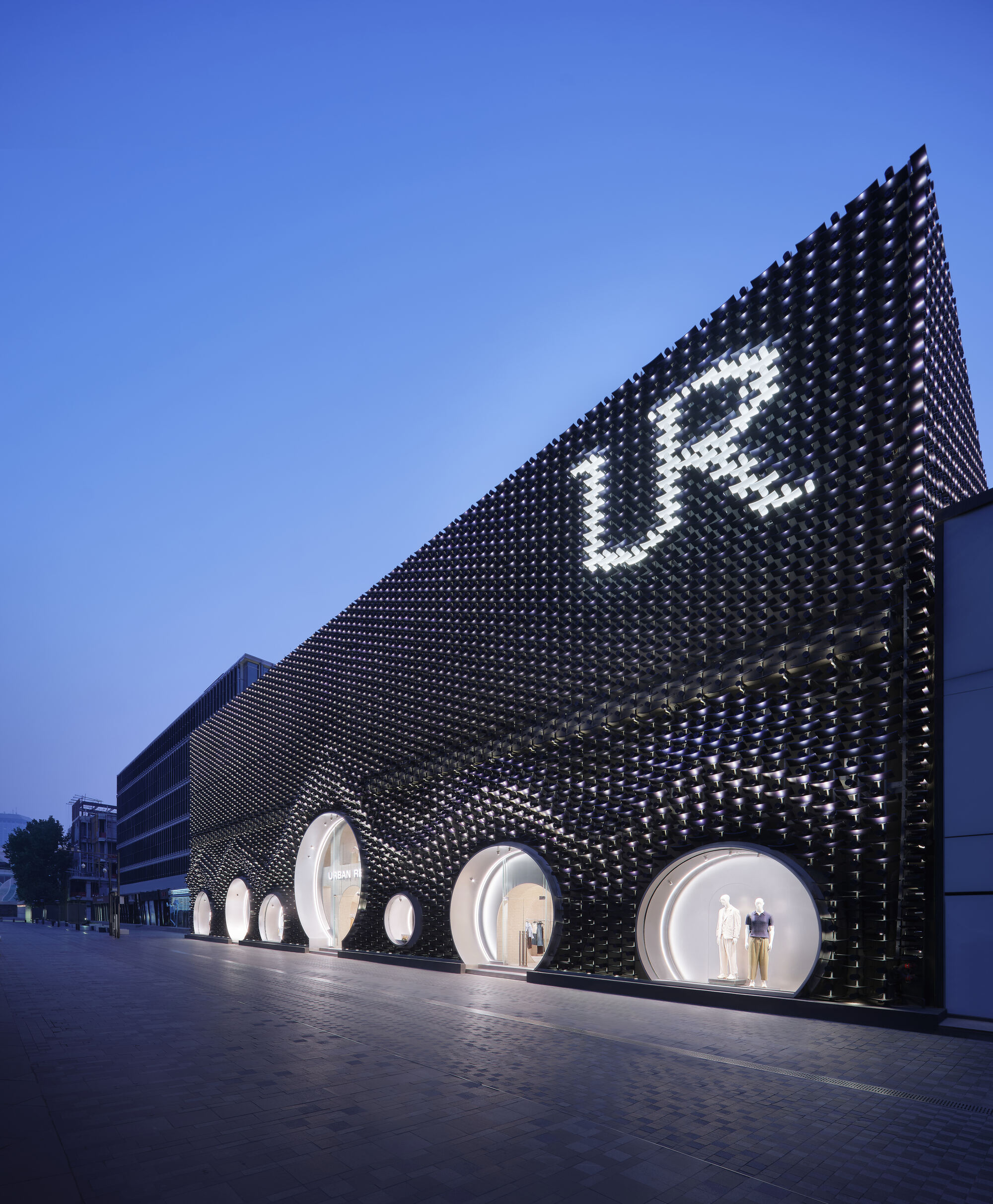 Long side facade of black peg cladding with round shop windows and a glowing sign under a deep blue evening sky