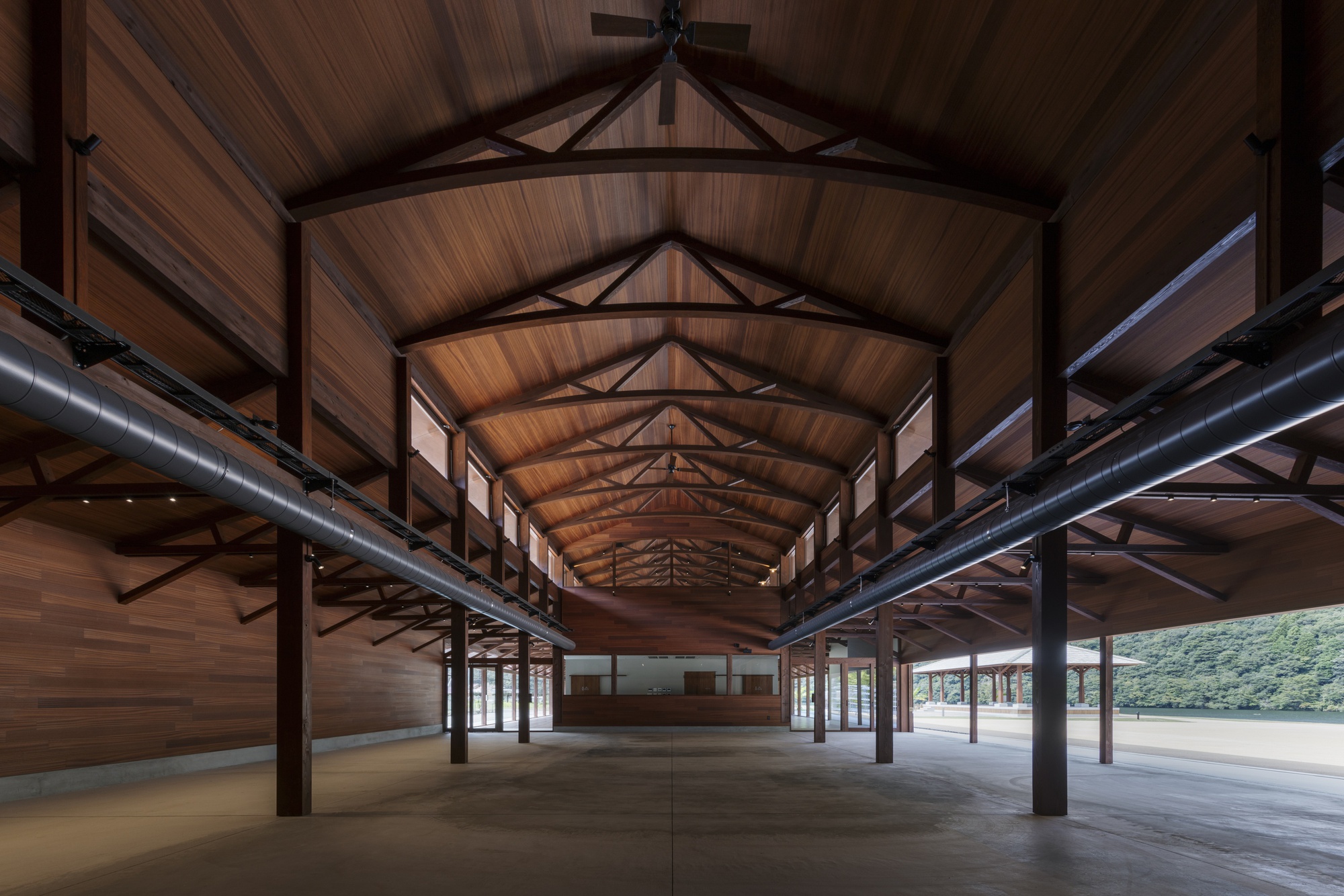 Open timber hall with arched trusses, dark columns and silver ducts running toward a far wall and green hillside